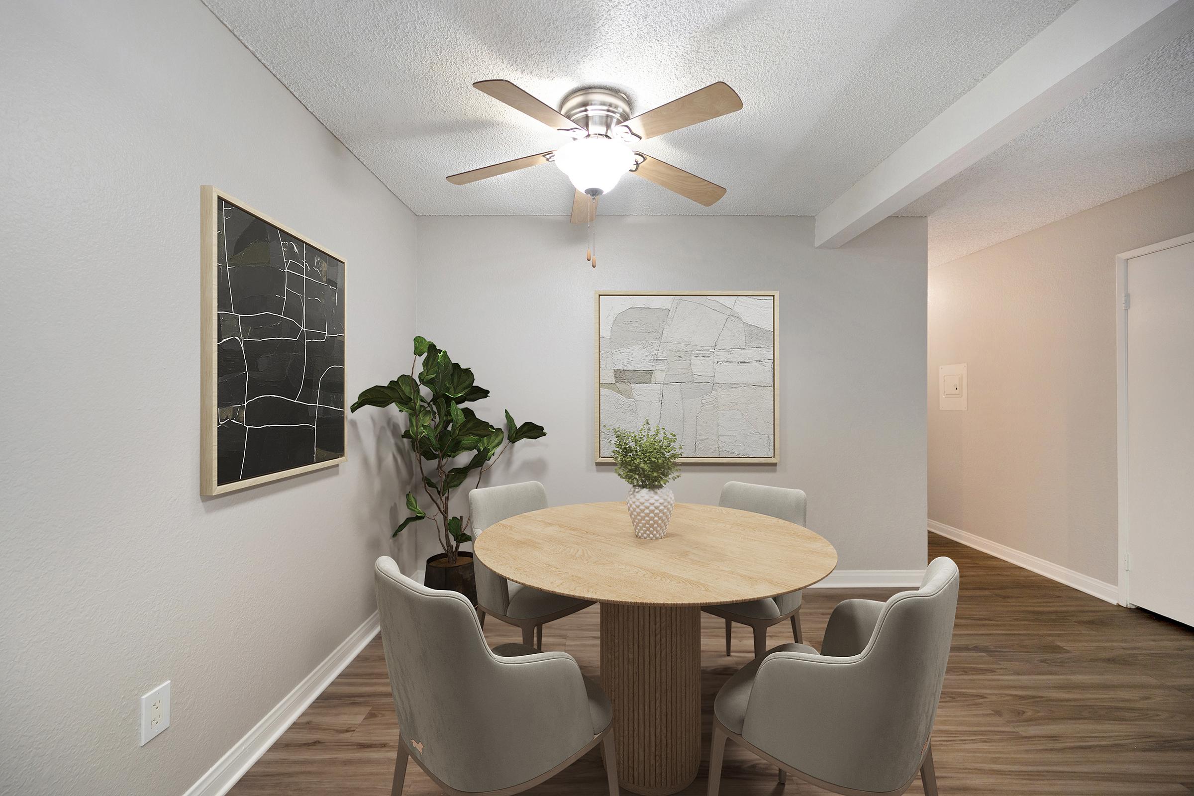 A cozy dining area featuring a round wooden table surrounded by four light-colored chairs. A ceiling fan hangs above. The walls are painted in a light gray tone, adorned with two framed artworks. A potted plant adds a touch of greenery to the corner, enhancing the inviting atmosphere.