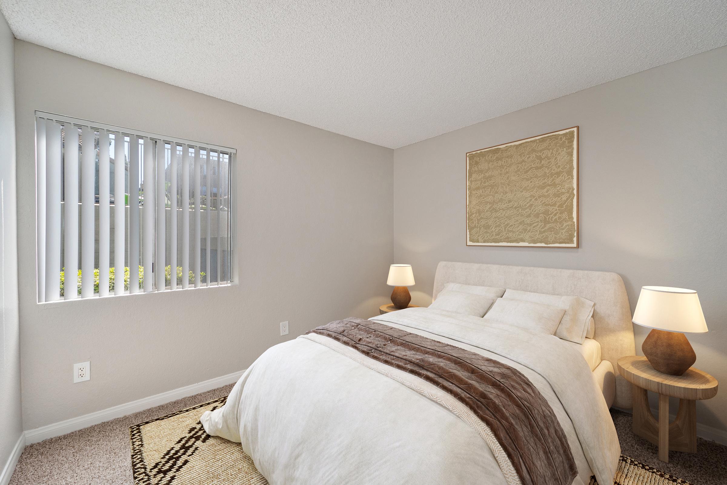 A cozy bedroom featuring a light-colored bed with a fluffy duvet, two bedside lamps, and a piece of wall art. The room has a window with vertical blinds, allowing natural light to illuminate the space. A textured area rug adds warmth to the floor.