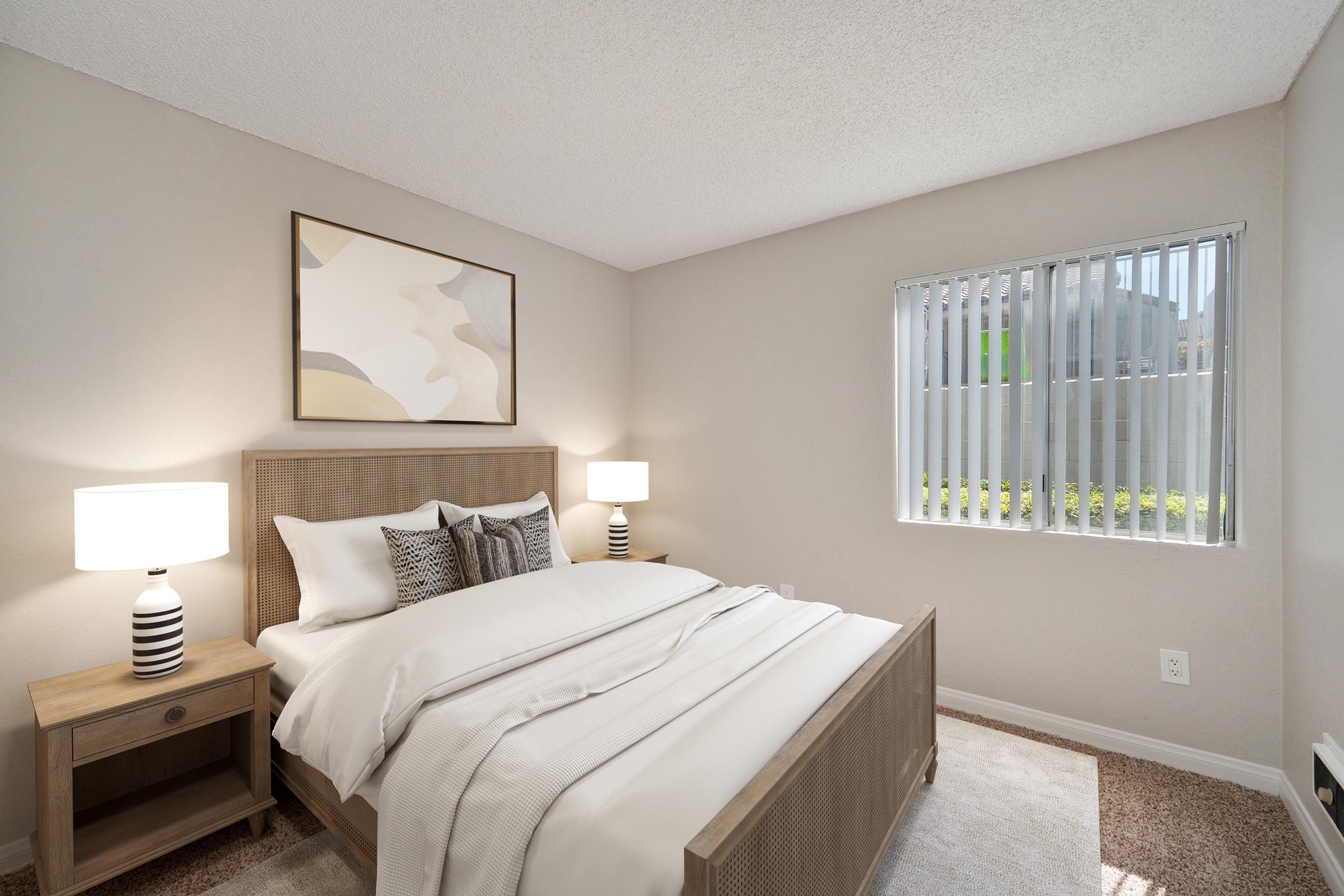 Cozy bedroom featuring a wooden bed frame with white bedding, two bedside lamps, and modern decor. A large window with vertical blinds allows natural light to fill the room, complemented by neutral wall colors and carpeting. A piece of abstract artwork adorns the wall above the bed.