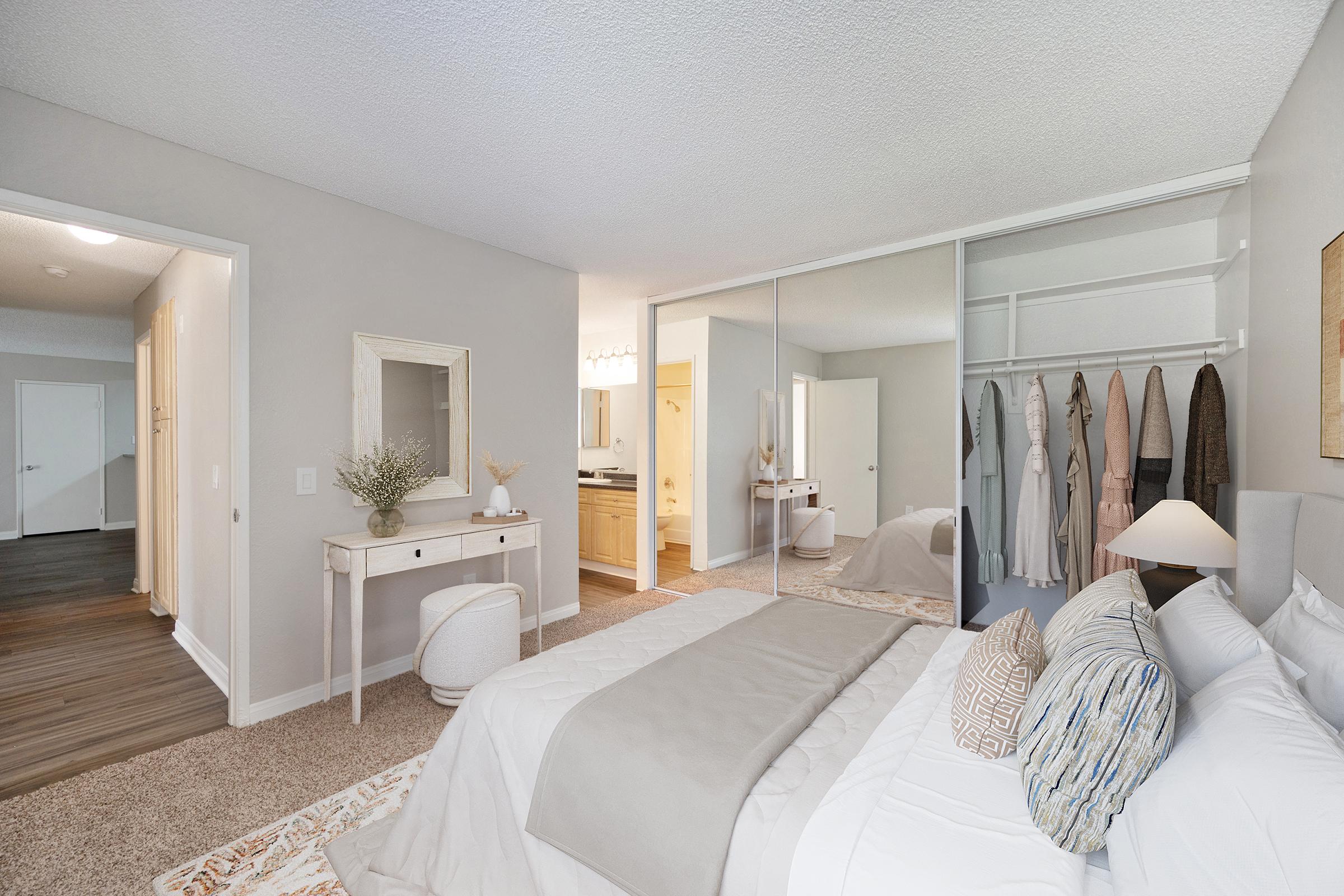 A modern bedroom featuring a large bed with neutral bedding, a stylish area rug, and a sleek nightstand. A full-length mirror and a closet with hanging clothes are visible, along with a small vanity table. Soft lighting creates a cozy atmosphere, and there are doors leading to a bathroom and hallway.