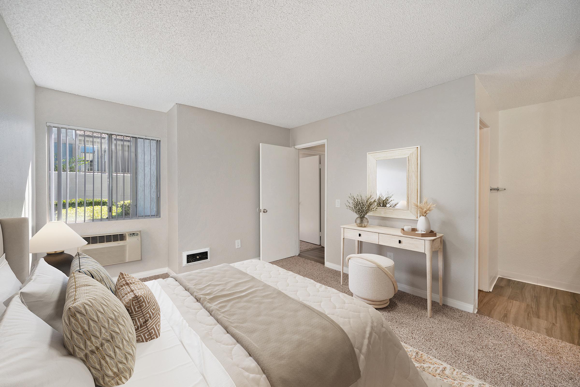 A cozy bedroom featuring a bed with decorative pillows, a wooden nightstand, and a mirror. The walls are painted in a light color, and there is a small air conditioning unit by the window. Natural light filters in, creating a warm atmosphere. A plush area rug and a small stool complement the space.