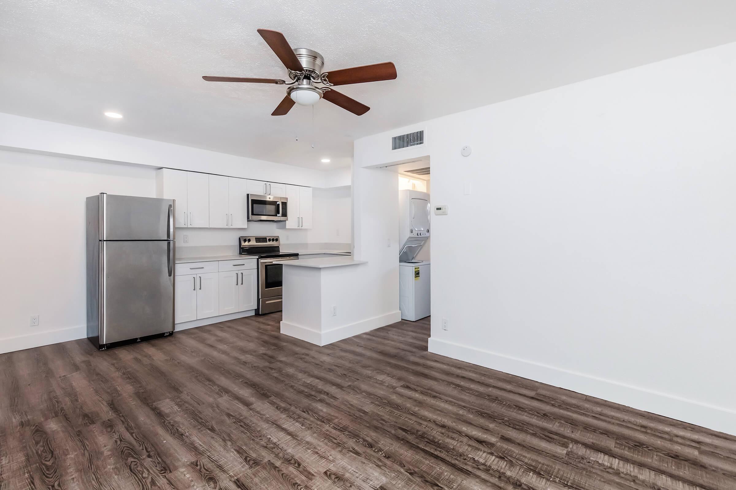 Modern kitchen and living area featuring stainless steel appliances, including a refrigerator and stove. The space has hardwood-style flooring, white cabinetry, and a ceiling fan. A laundry area is accessible in the background, with bright, neutral-colored walls enhancing the open layout.