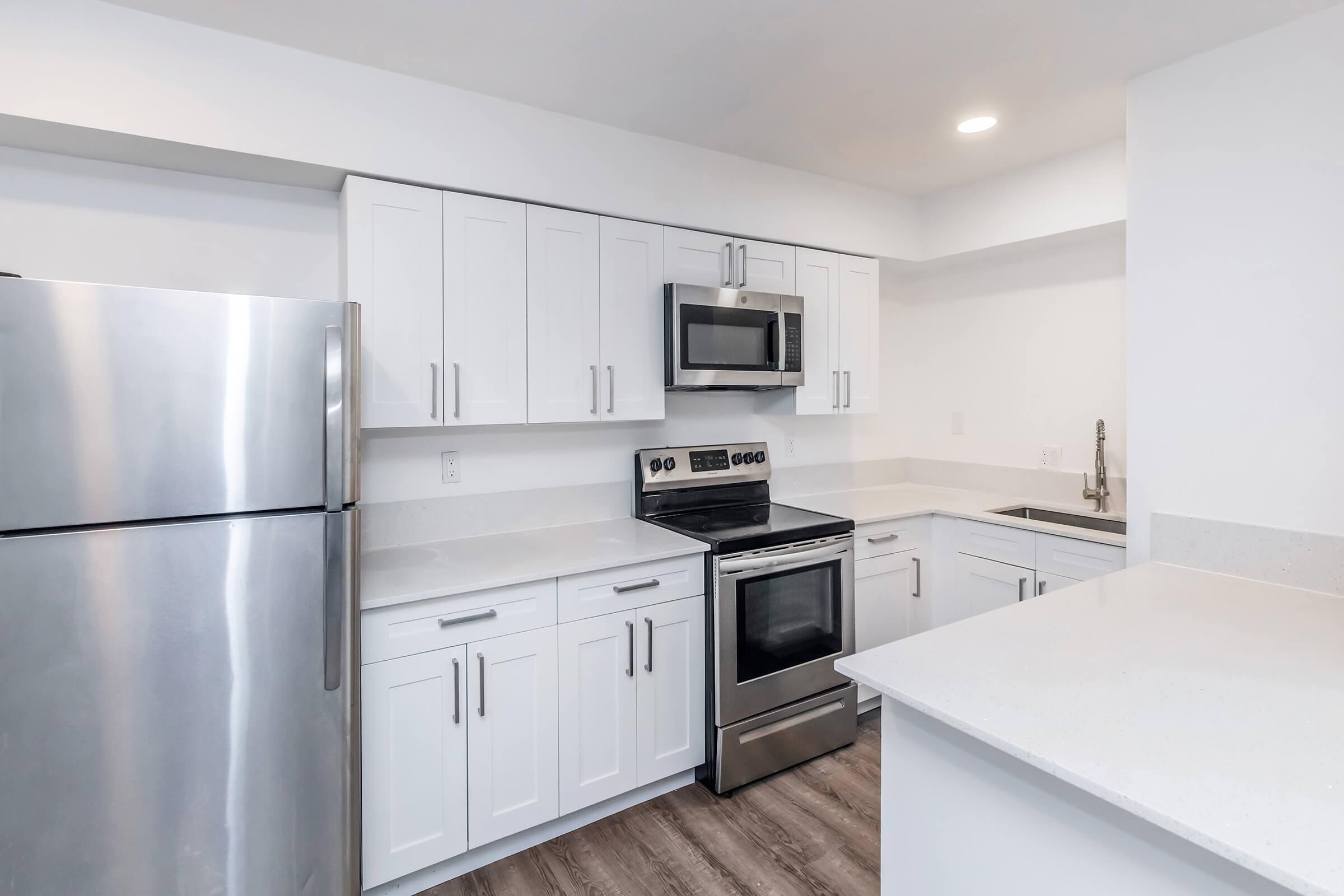 Modern kitchen with white cabinetry, stainless steel appliances including a refrigerator, oven, and microwave, and a light-colored countertop. The space is bright and clean, featuring light wood-like flooring and a simple, functional design.