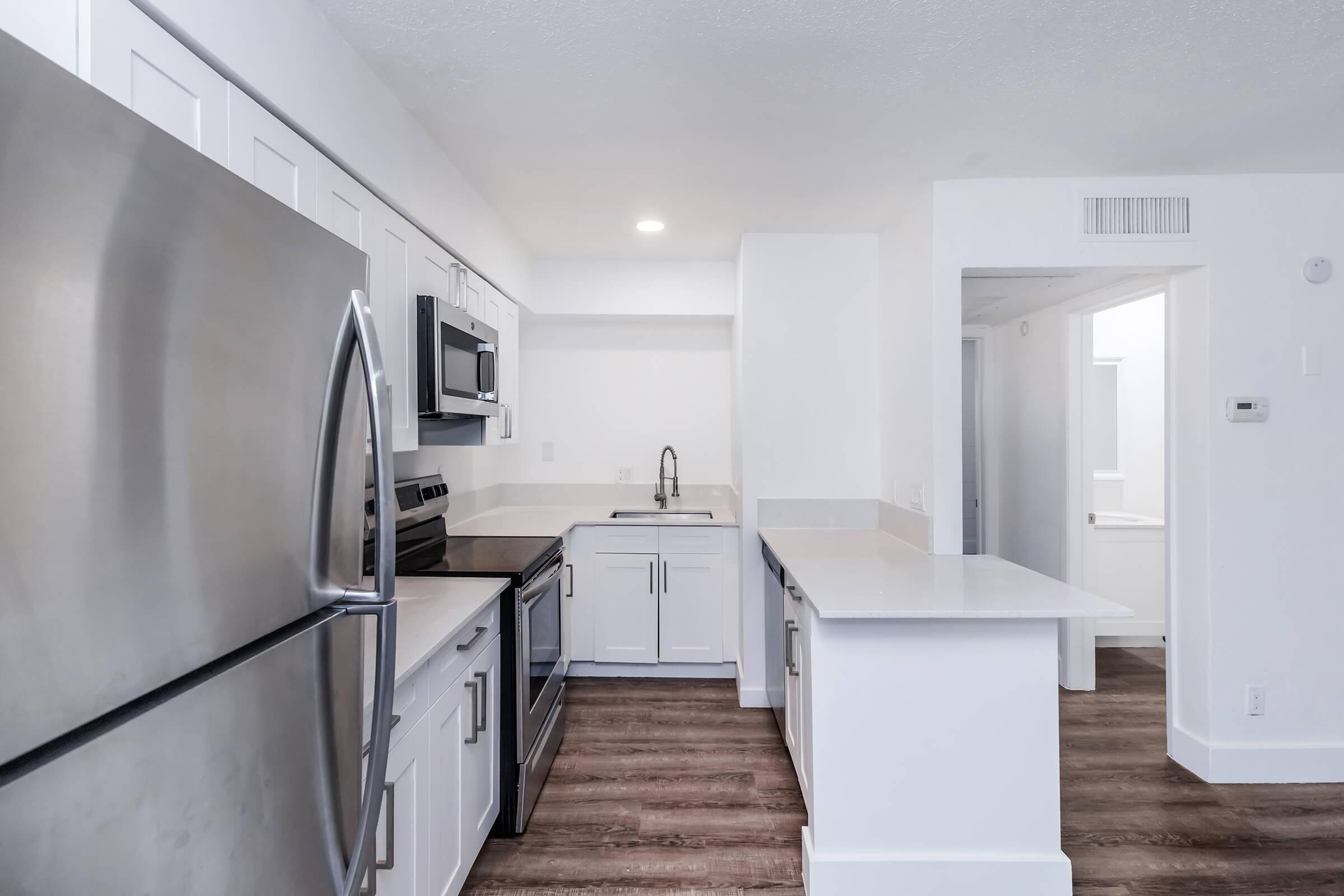 Modern kitchen featuring stainless steel appliances, including a refrigerator, oven, and microwave. The kitchen is characterized by white cabinetry, a light countertop, and wood-like flooring. A sink is installed with a sleek faucet, and there is a bright, open layout leading to adjacent rooms.