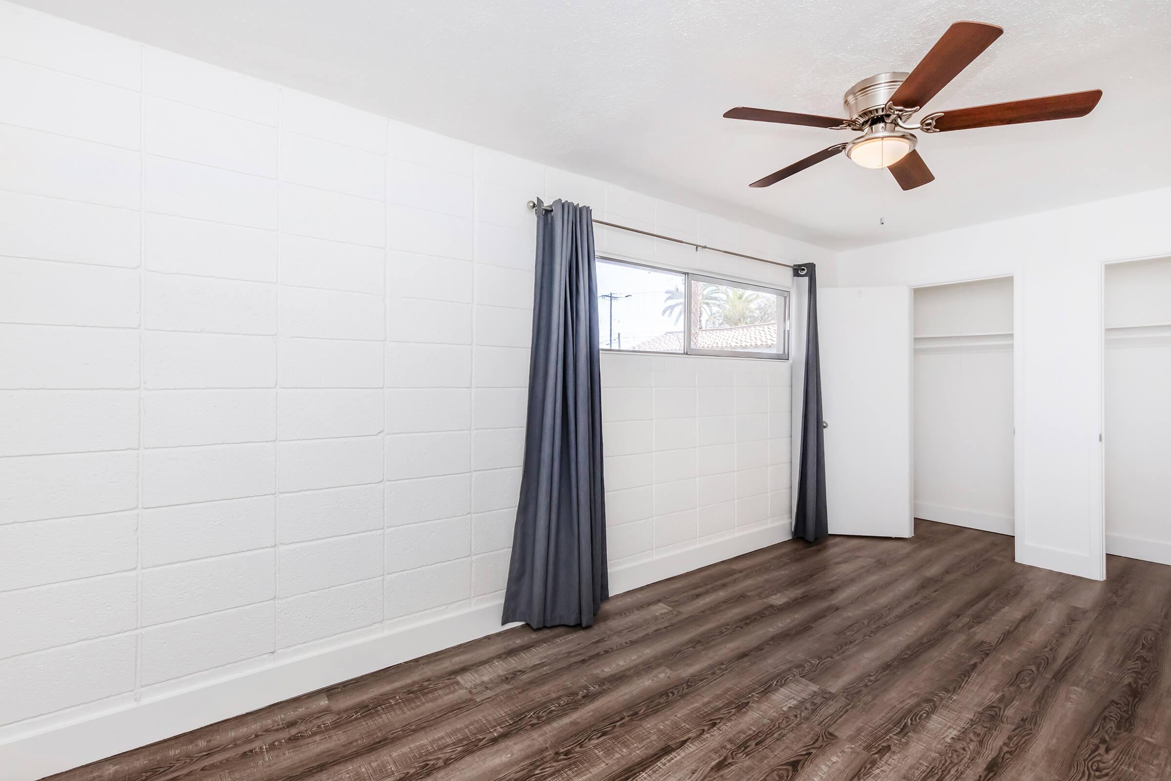 A brightly lit room featuring white walls with a textured finish, a ceiling fan with wooden blades, and large window covered with gray curtains. The floor is made of modern dark wood laminate, and an open closet is visible on the right side, creating a clean and spacious atmosphere.