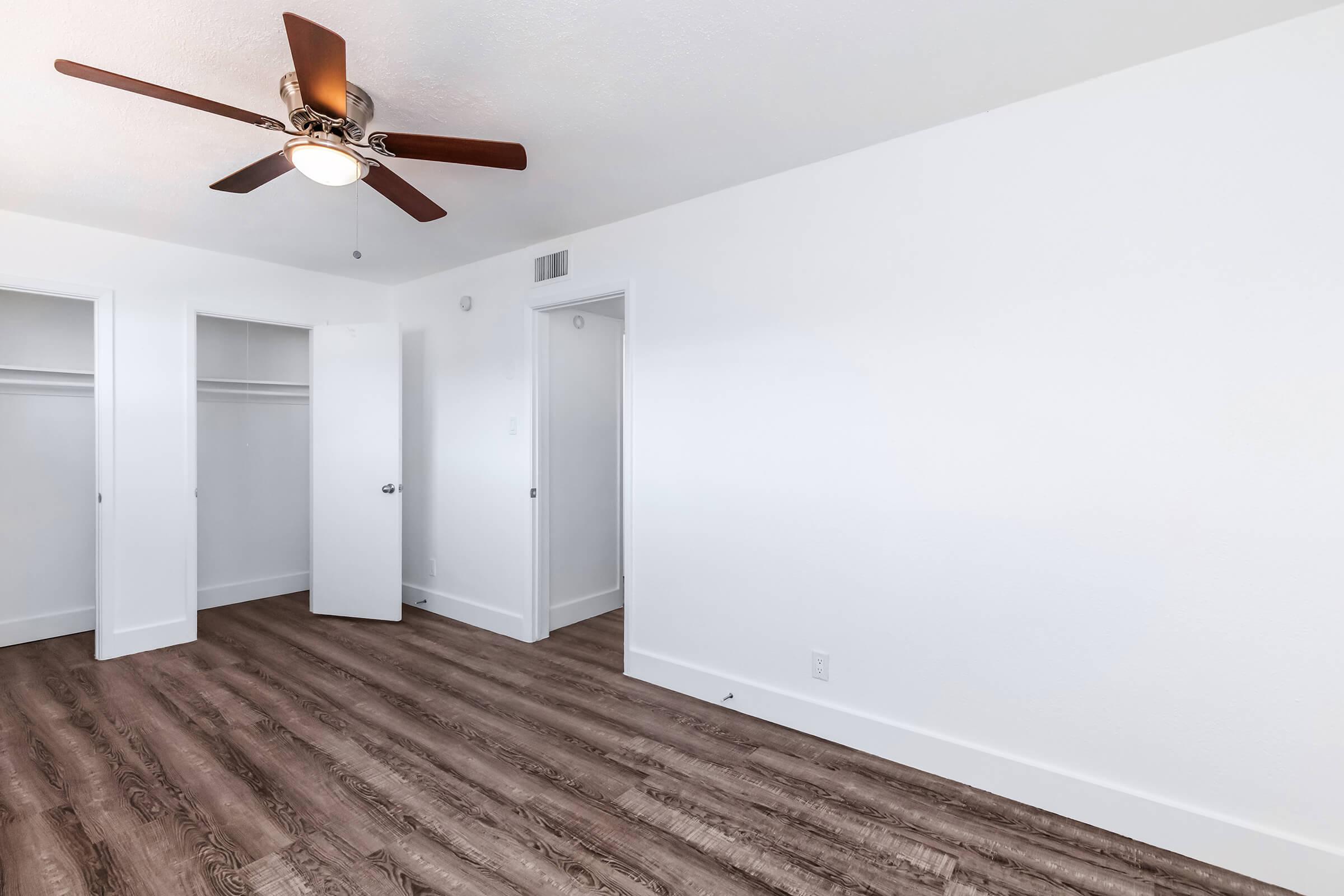 A well-lit room with white walls and a ceiling fan. The flooring is a light brown wood laminate. There are two white closet doors on the left, and a plain wall on the right, leading to a doorway. The room is empty and spacious, conveying a clean and modern aesthetic.