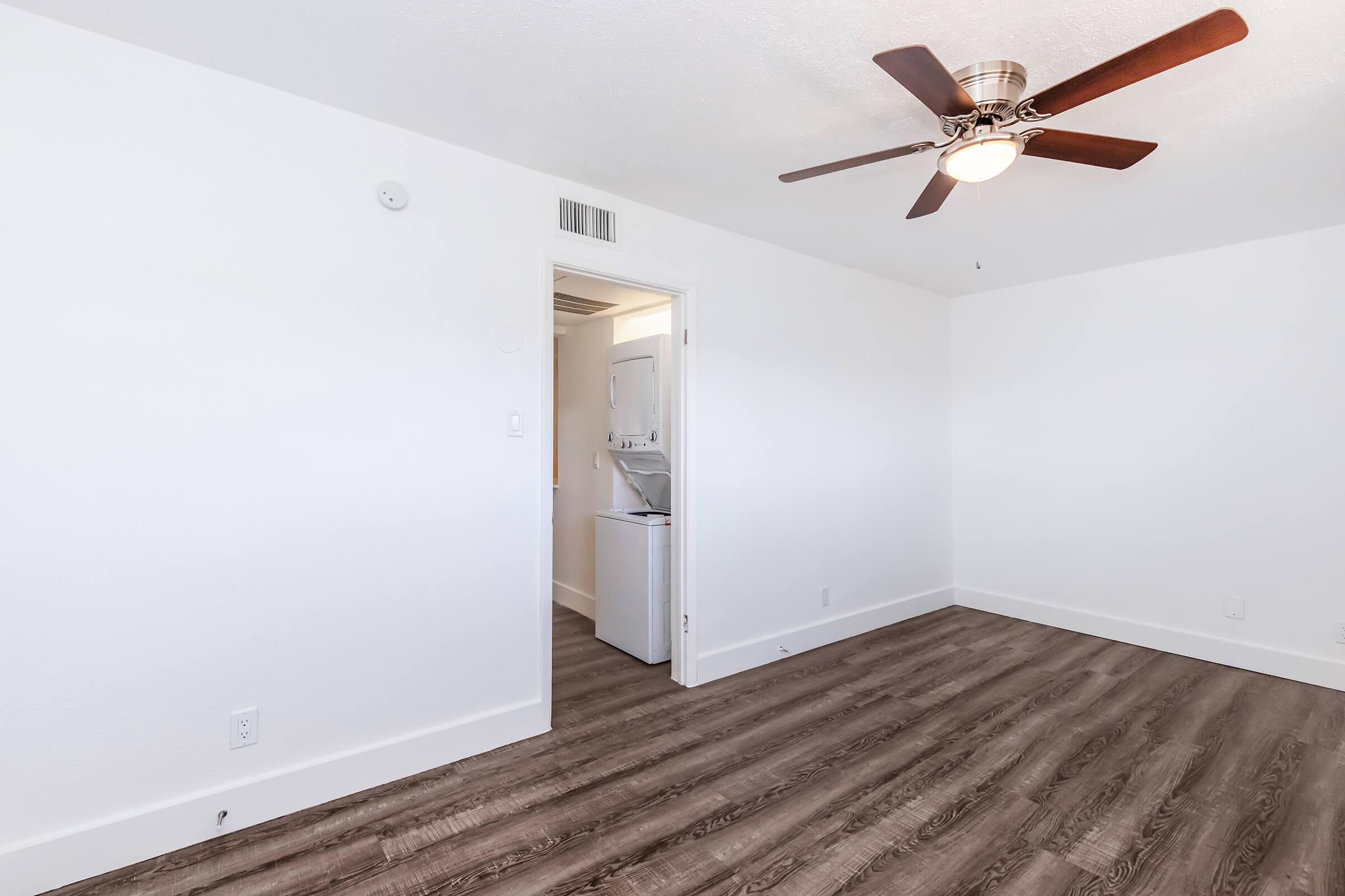 A bright, empty room featuring light-colored walls and a ceiling fan. On the left, there's an open door leading to a laundry area with a washer and dryer. The floor is dark wood, adding warmth to the space. Perfect for a cozy living environment.
