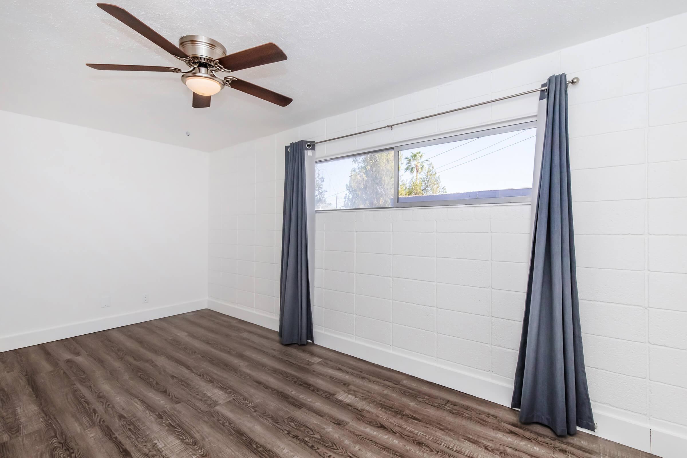 A spacious room with light wood flooring, featuring a ceiling fan and a large window covered by dark curtains. The walls are painted white, creating a bright and airy atmosphere. The room is empty, allowing for various furniture arrangements.