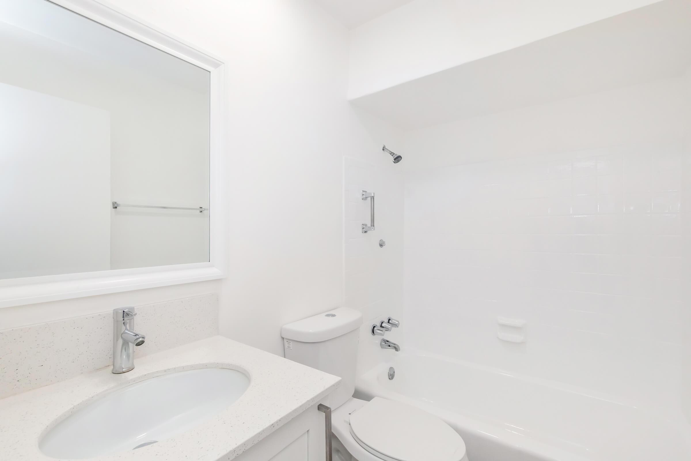 A clean, modern bathroom featuring a white sink with a faucet, a toilet, and a bathtub with a shower. The walls and tiles are bright white, creating a spacious and airy feel. A large mirror above the sink adds to the light atmosphere.