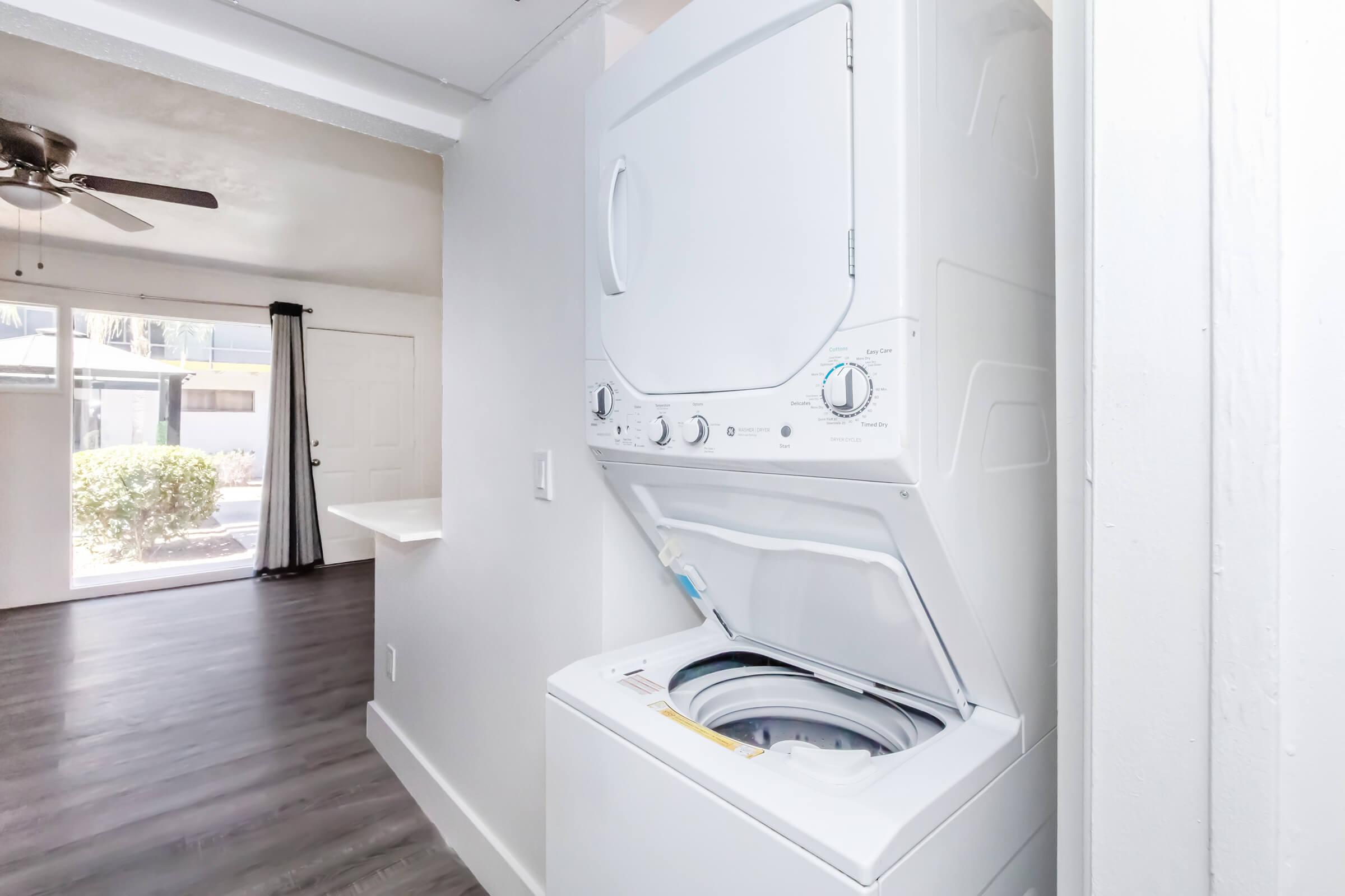 A stacked washer and dryer unit in a modern, light-colored laundry space. The room features a ceiling fan and a view of a living area with a large window. There are gray laminate floors and white walls, creating a clean and bright atmosphere.