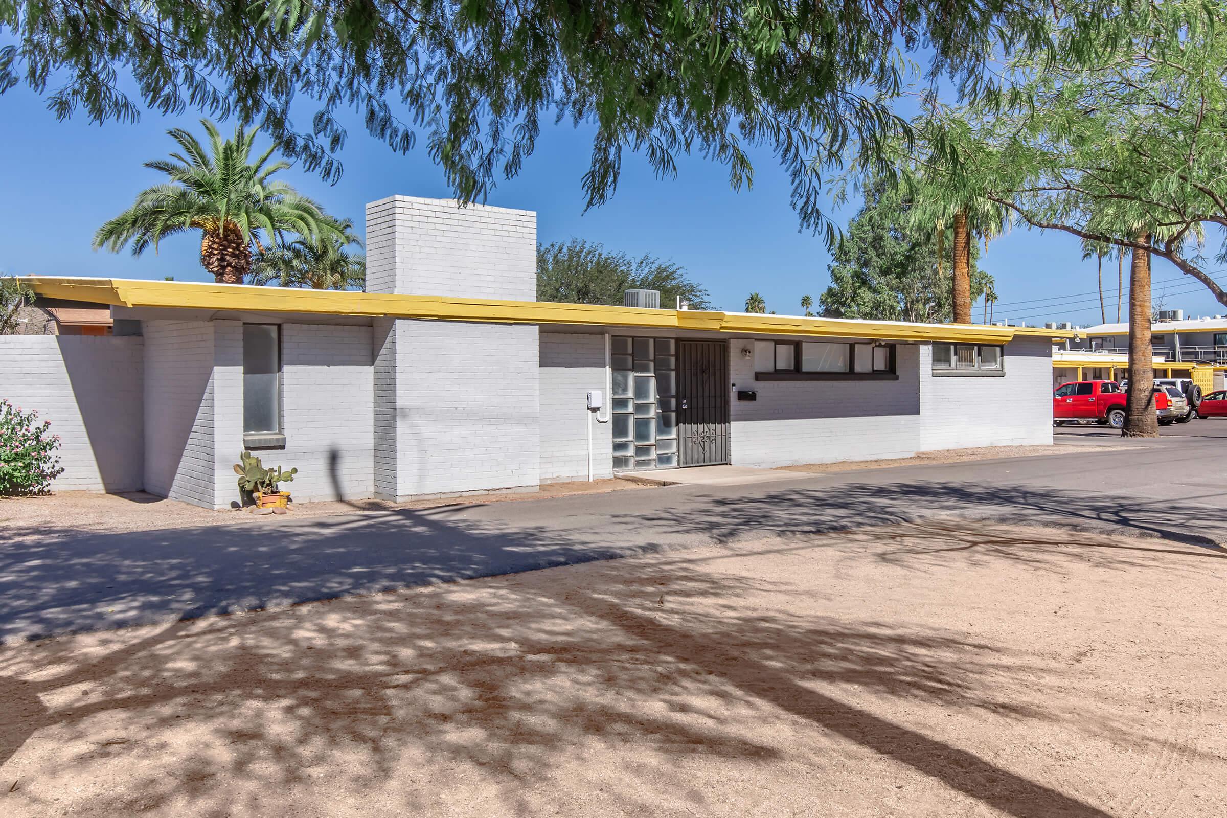 A single-story, mid-century modern home with a yellow overhang and gray brick exterior. The property features palm trees in the background, a gravel front yard, and a simple, clean design typical of desert architecture. The street is lined with similar homes, creating a serene neighborhood atmosphere.
