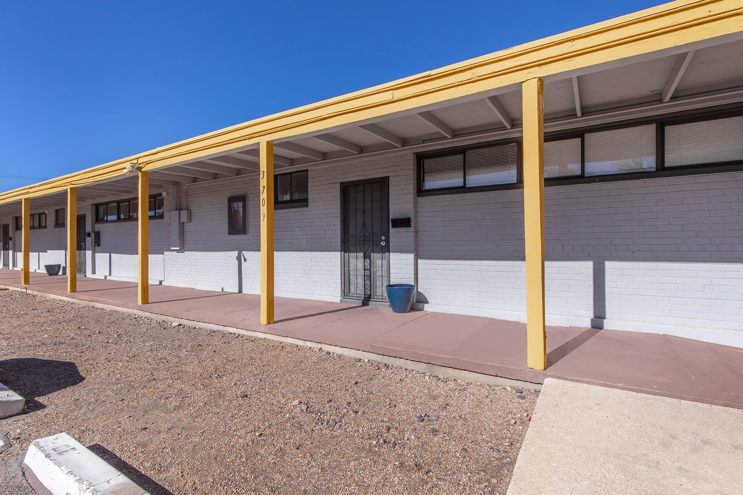A well-lit exterior view of a single-story building with a yellow overhang. The structure features a series of windows and a central door with a security gate. The surrounding area has gravel landscaping, and there is a blue planter near the entrance. The sky is clear and blue.