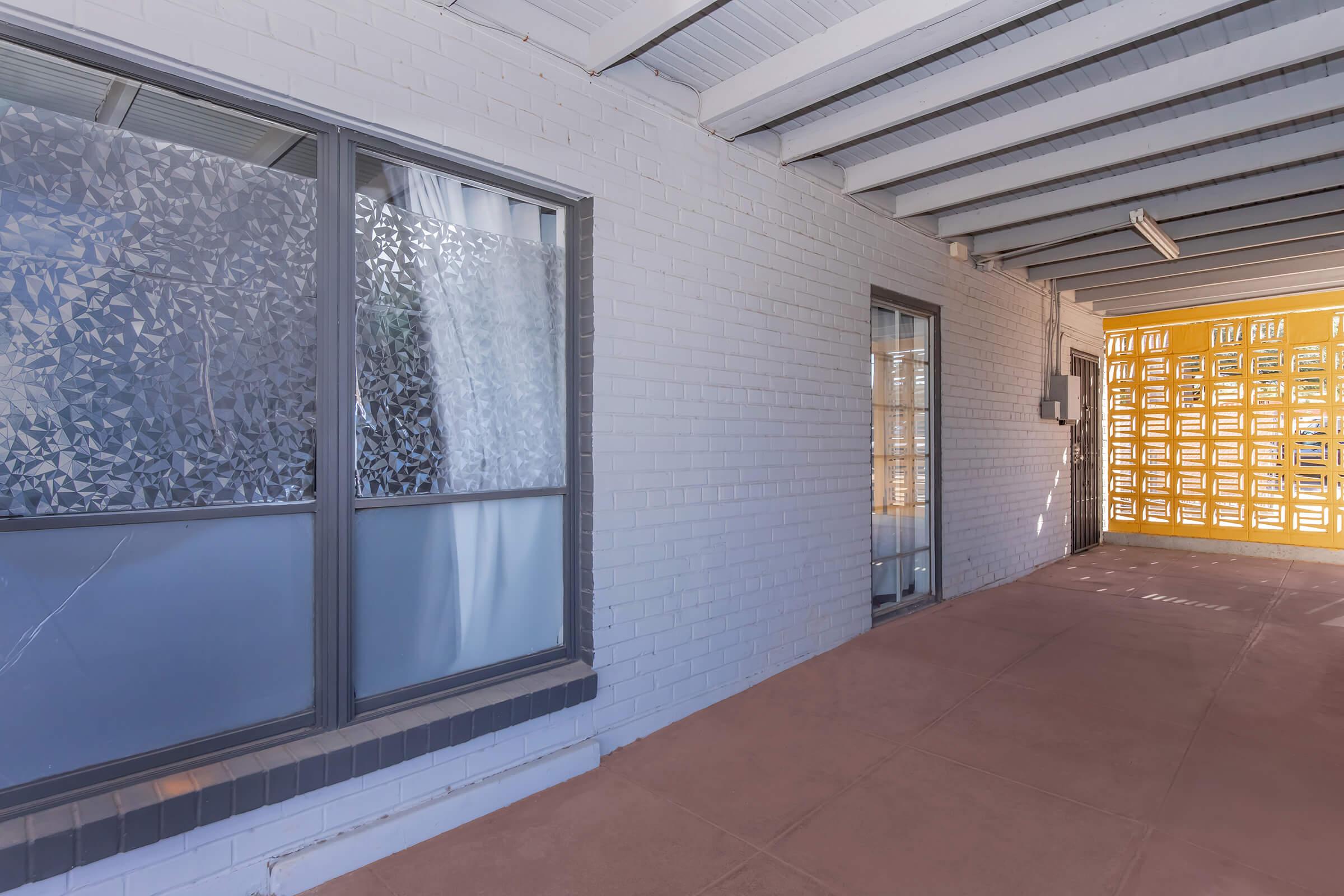A view of a corridor with white brick walls and windows. One window has a patterned design, while the other is clear. The floor is a light-colored tile, and there is a wall with a grid pattern on the right side. Natural light illuminates the space, creating a bright and airy atmosphere.