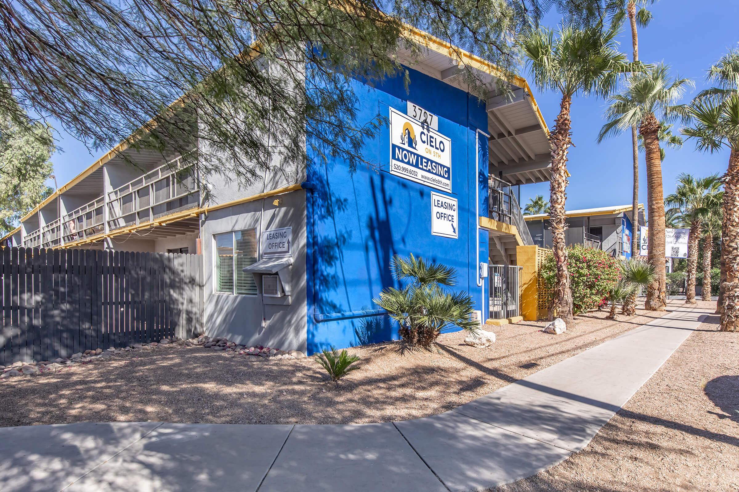 A colorful two-story building with a blue and yellow facade, surrounded by palm trees and desert landscaping. A sign on the building indicates it is a leasing office, with a pathway leading from the sidewalk to the entrance. The scene conveys a sunny, inviting atmosphere.