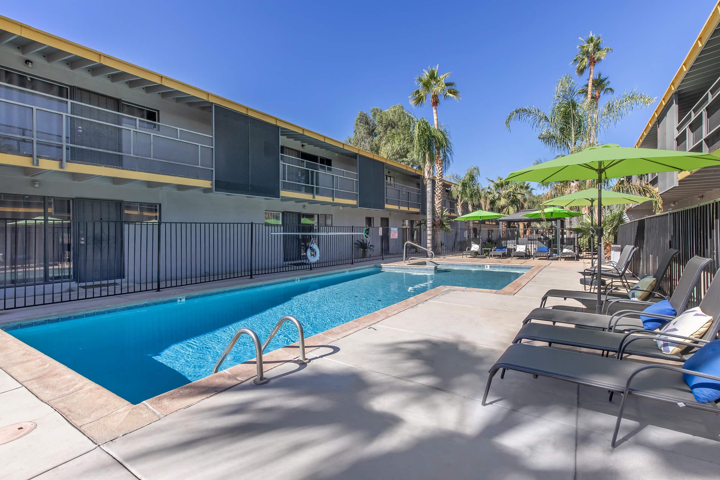 Swimming pool surrounded by lounge chairs and bright green umbrellas, with palm trees and a sunny sky in the background. Two-story buildings with balconies line one side of the pool area, creating a relaxing atmosphere for leisure and enjoyment.