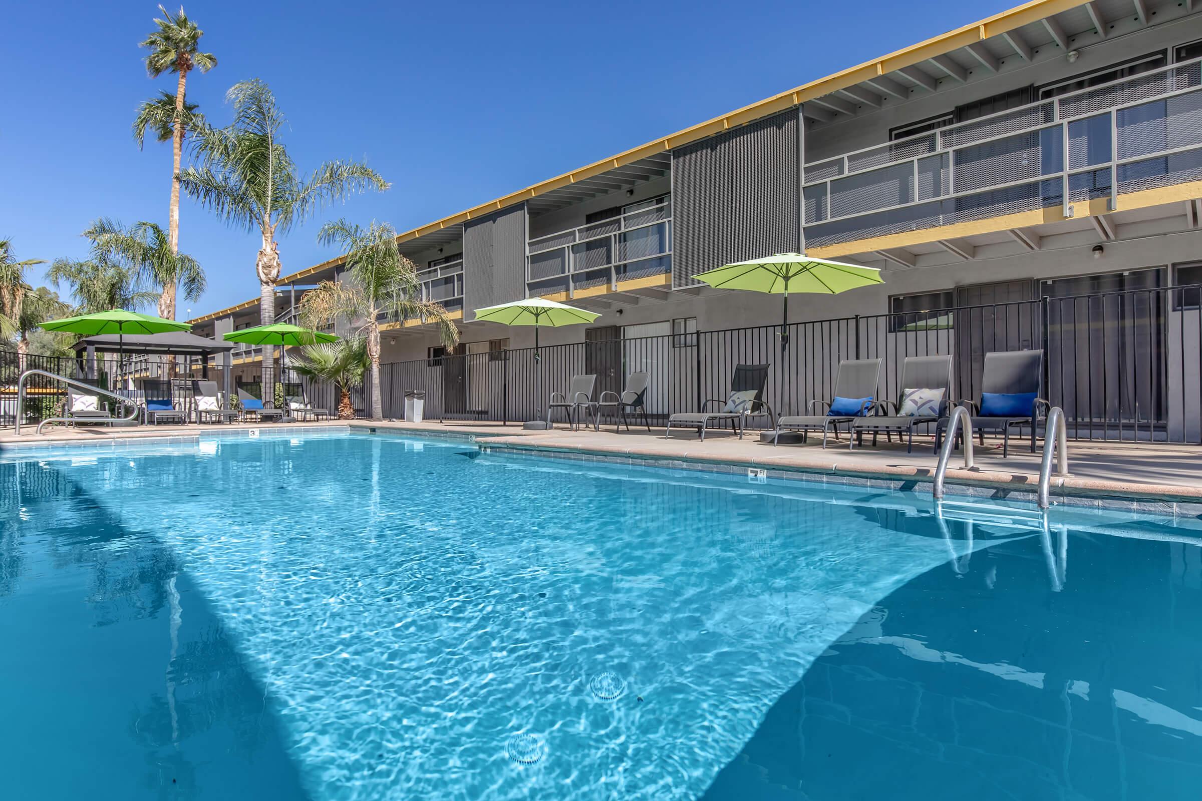 A clear blue swimming pool with lounge chairs and green umbrellas, surrounded by palm trees. In the background, there are two-story buildings with balconies under a bright blue sky. The scene conveys a relaxing and inviting atmosphere, ideal for leisure and enjoyment.