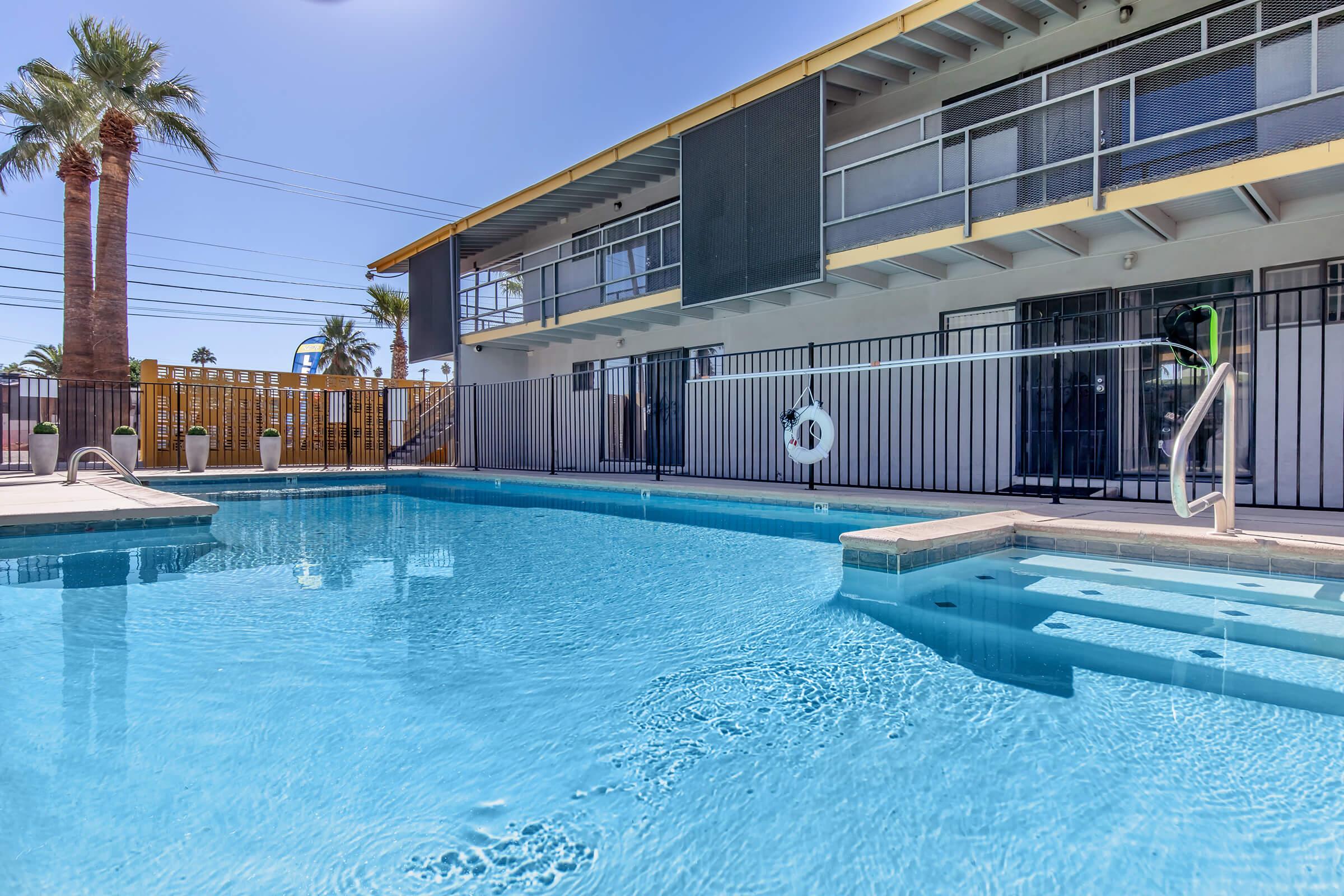 A clear blue swimming pool surrounded by a pool deck, with a two-story building in the background featuring balconies. Palm trees are visible nearby, and a lifebuoy hangs on the poolside railing. The scene is set under a bright, sunny sky.