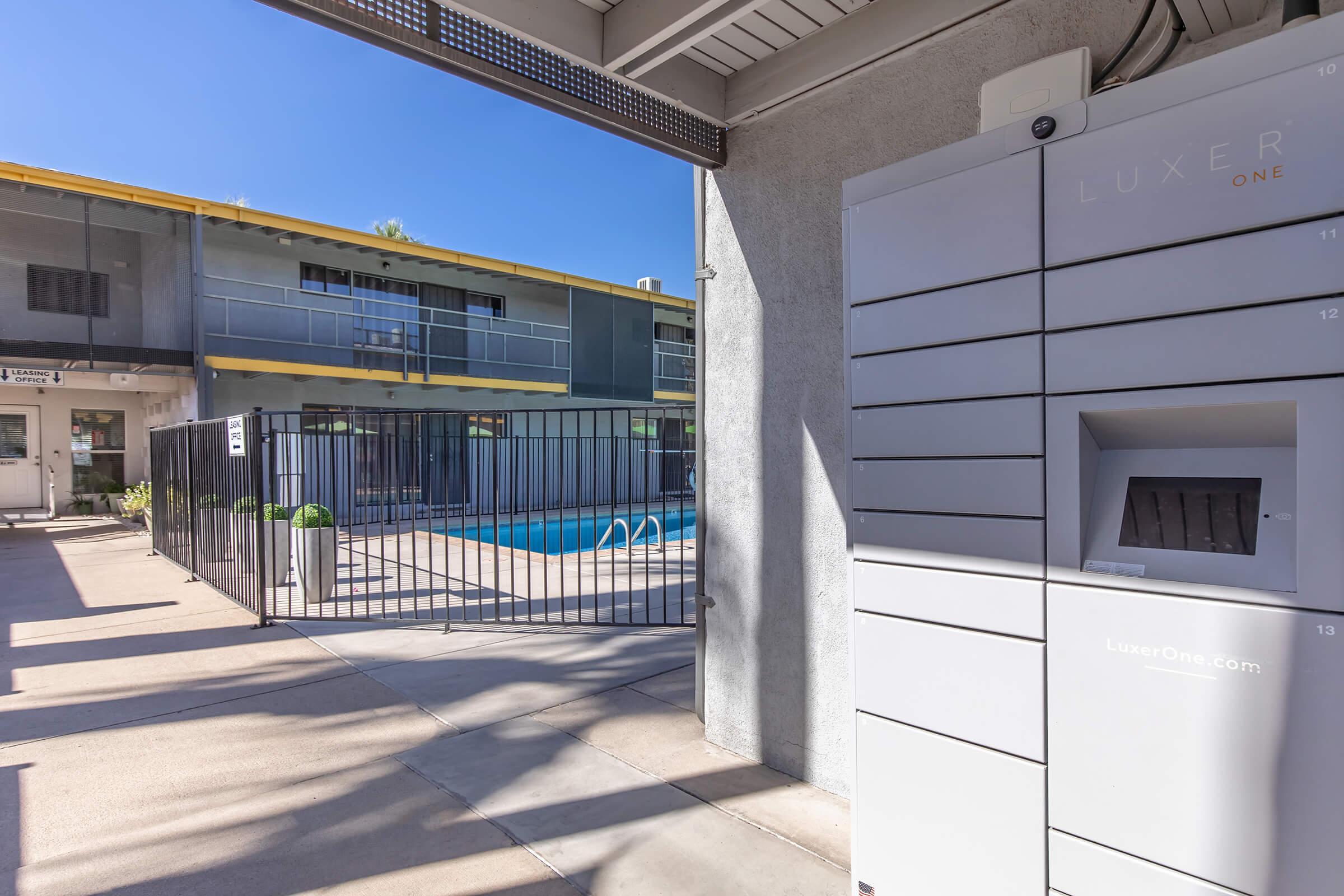 A view of an apartment complex featuring a swimming pool, a secure parcel locker system labeled "Luxer One," and a walkway leading to the entrance of a building. The setting is bright and sunny, with modern architecture and minimal landscaping.