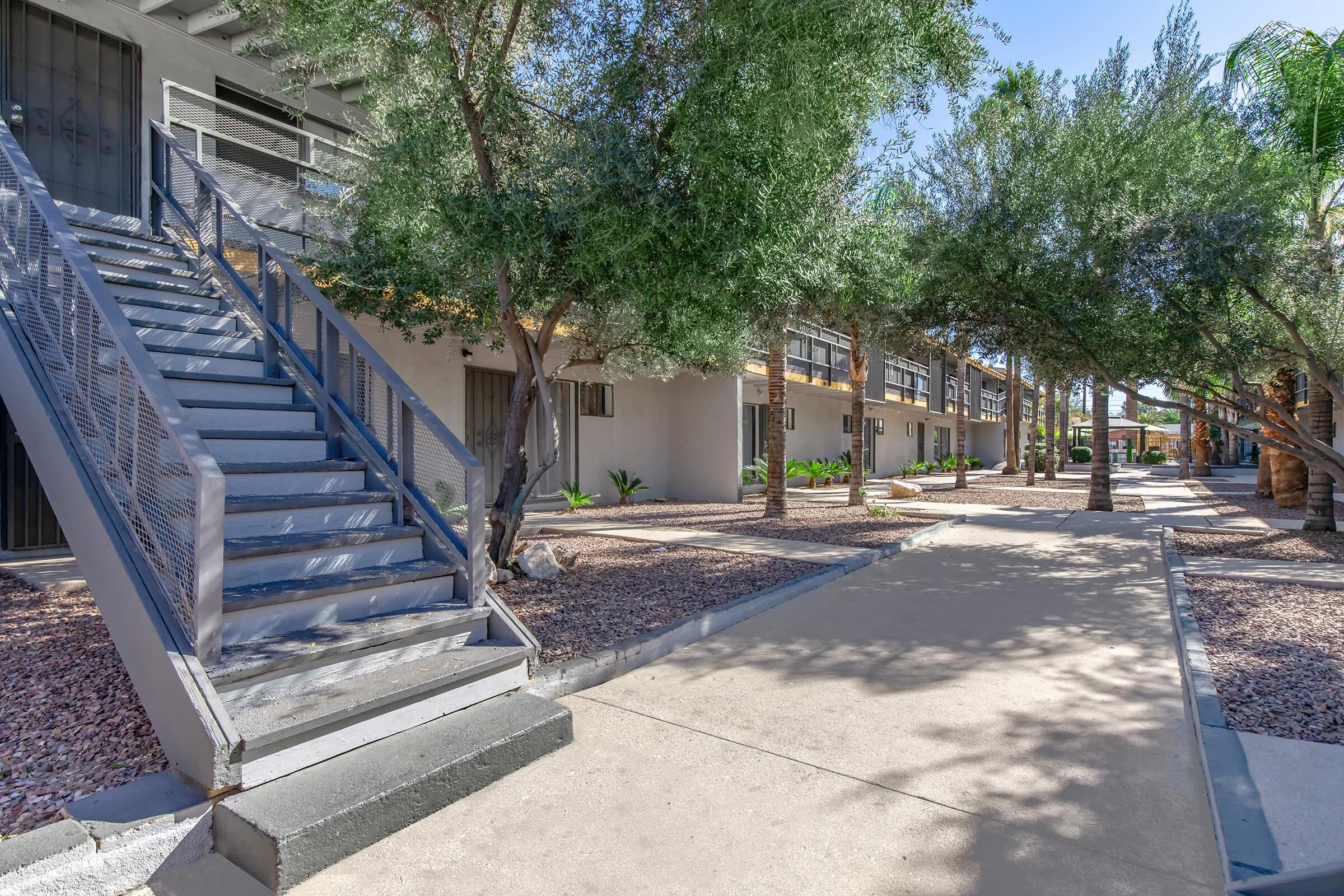 A modern outdoor apartment complex featuring a staircase leading to upper units, lined with lush trees and shrubs. The pathway is paved, connecting different sections of the building with well-maintained landscaping under a clear blue sky.