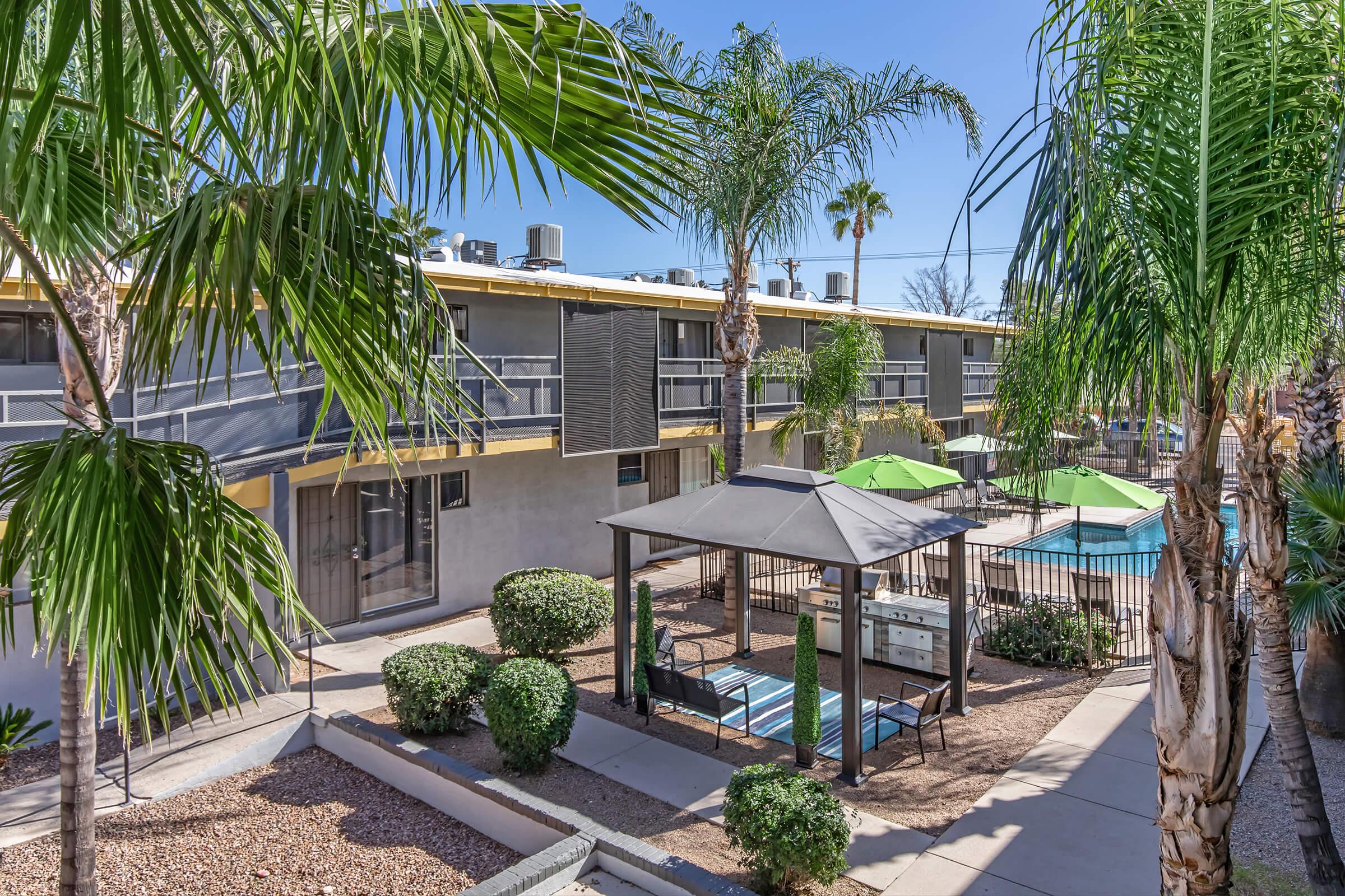 A view of a landscaped courtyard featuring palm trees, a gazebo, and a fenced swimming pool area. Surrounding buildings have balconies, and patio furniture is visible. The scene conveys a sunny, inviting atmosphere typical of a resort or apartment complex.