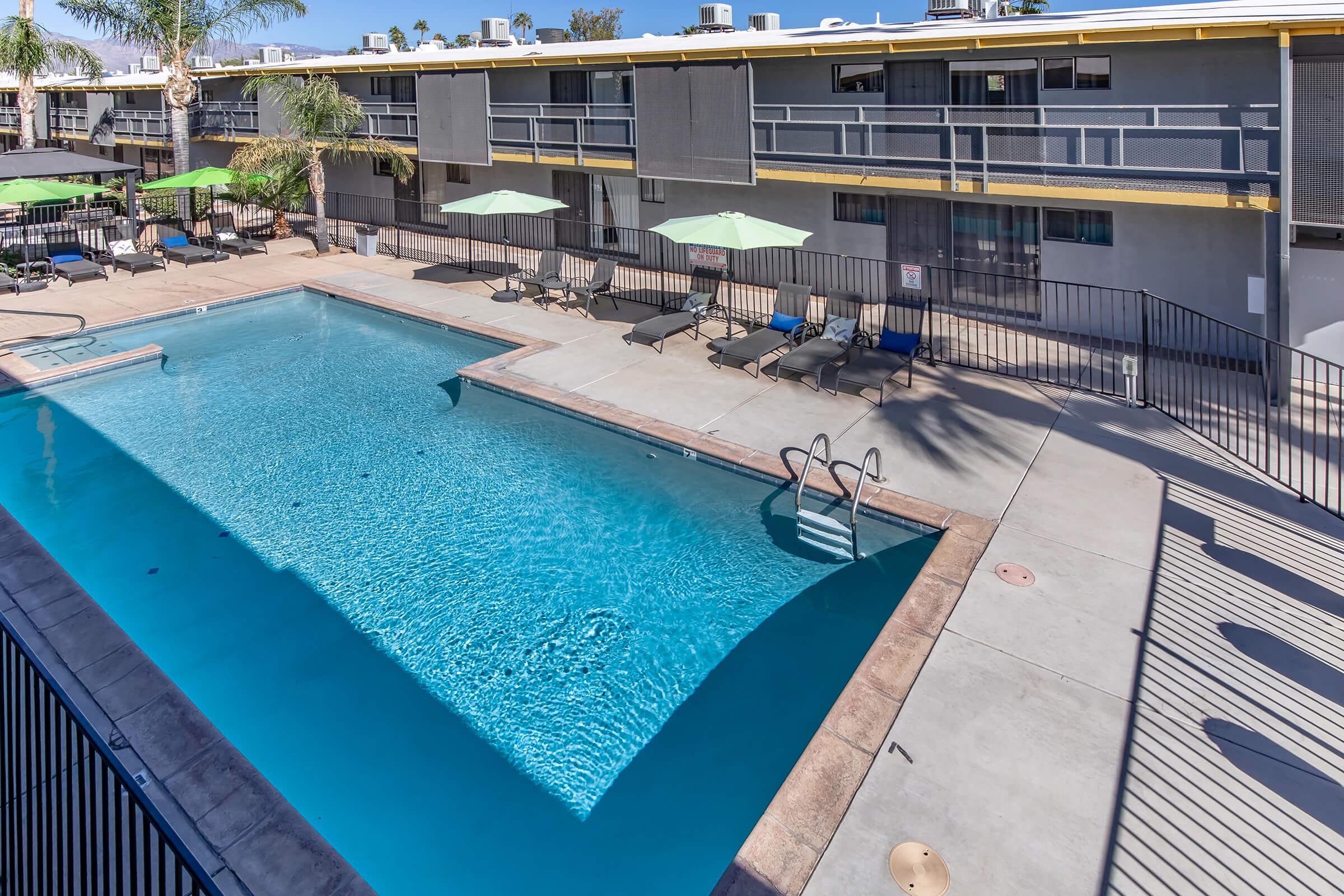 A clear blue swimming pool surrounded by lounge chairs and green umbrellas, with a two-story building in the background. The scene is set in a sunny outdoor area, showcasing a clean and inviting recreational space.
