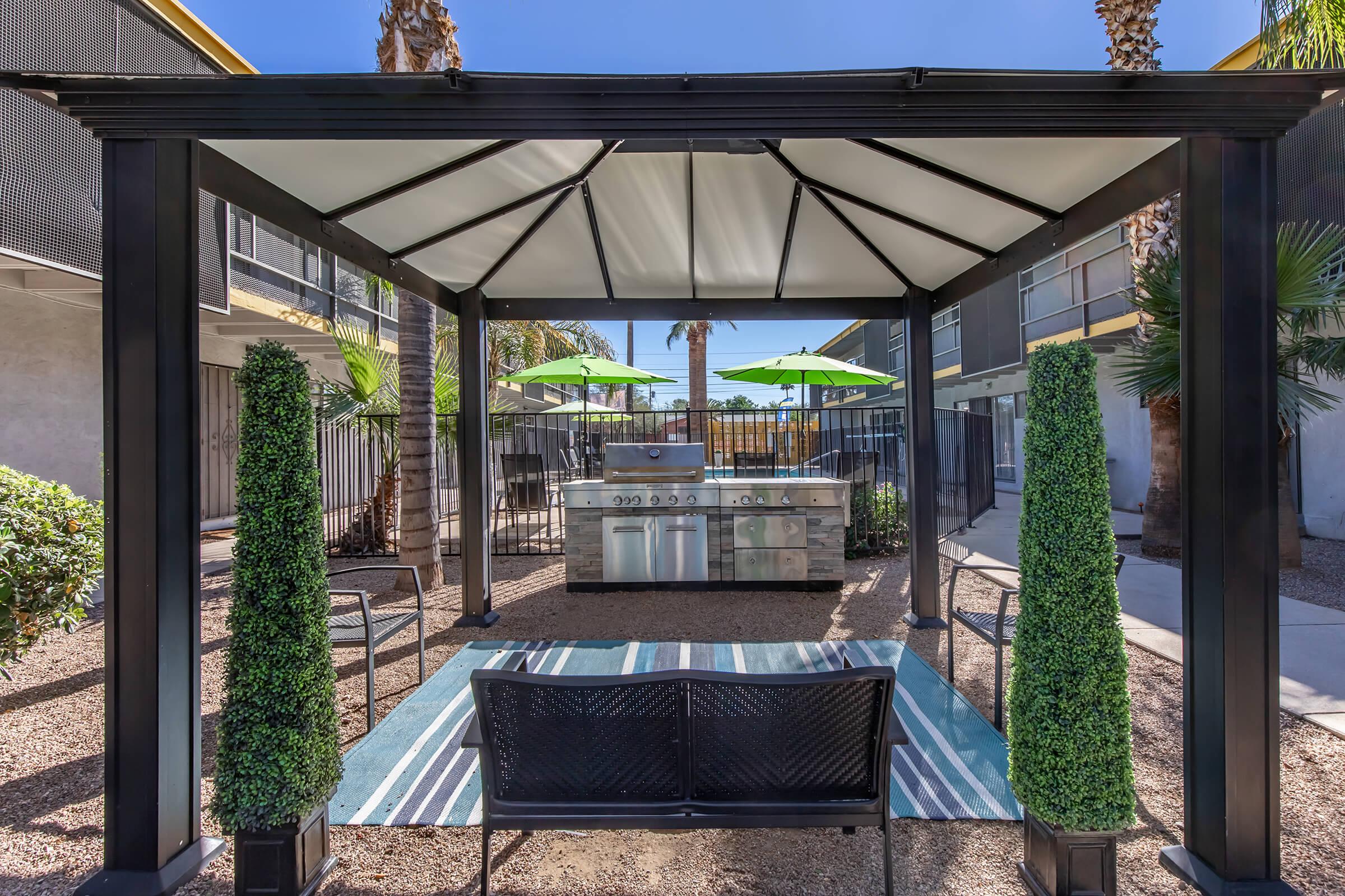 A patio area featuring a covered grill station with a stainless steel barbecue, surrounded by vibrant green umbrellas and neatly trimmed hedges. A black bench sits on a blue-striped rug in the foreground, with a backdrop of palm trees and an apartment building. Clear blue sky overhead.
