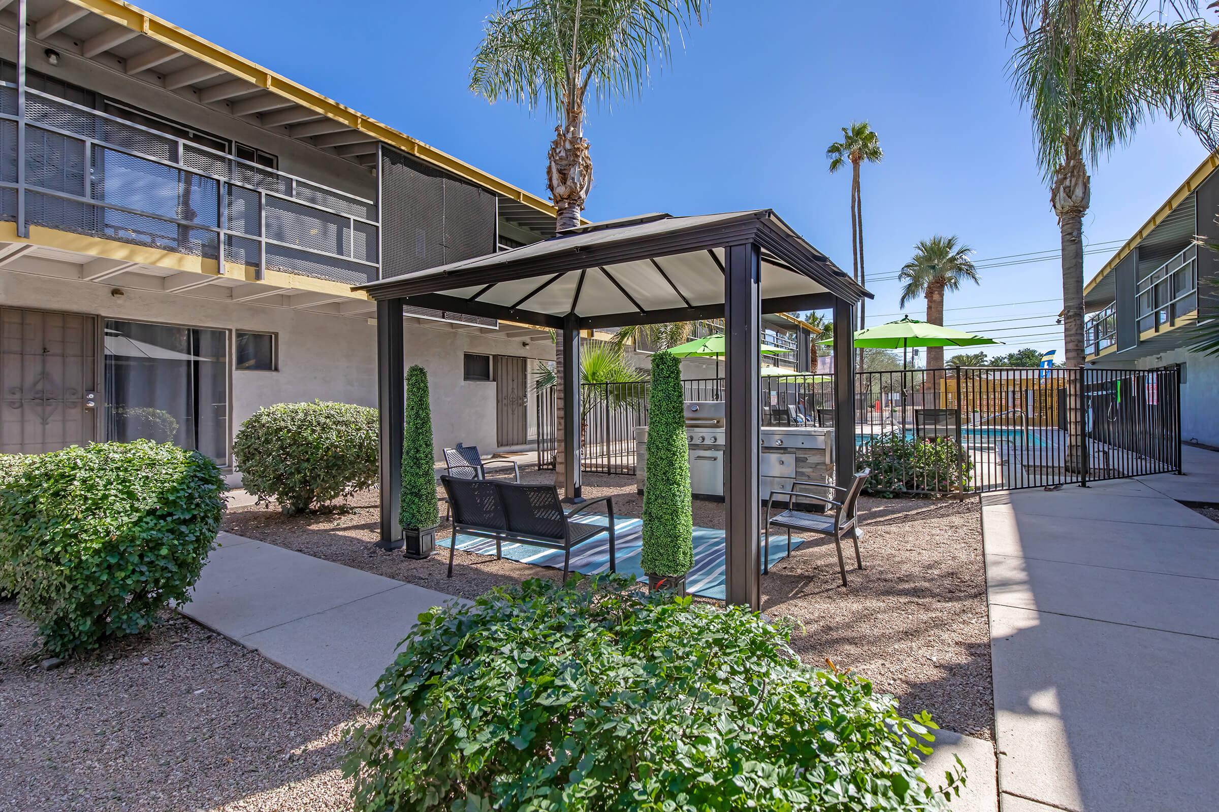 A landscaped courtyard featuring a gazebo with seating, surrounded by greenery and palm trees. In the background, there's a pool area enclosed by a fence and shaded by bright green umbrellas, alongside a building with a modern architectural design. Clear blue skies overhead.