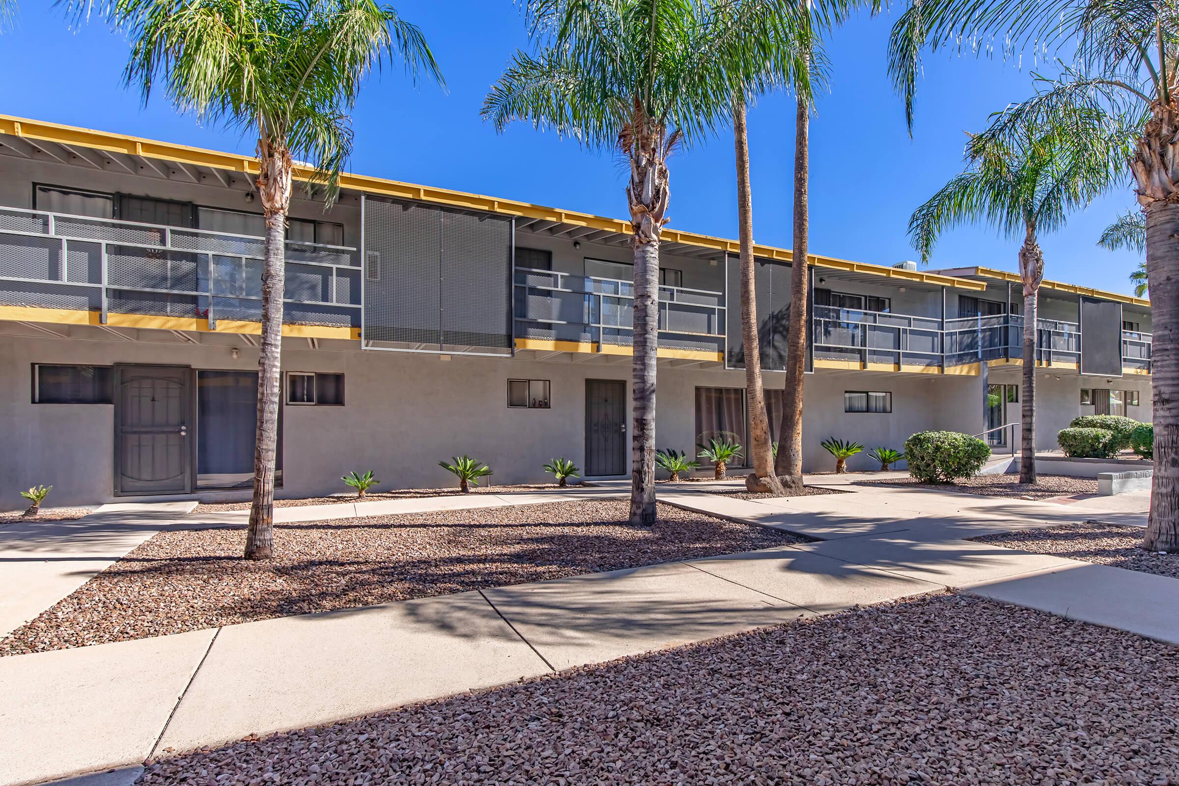 A modern apartment complex featuring multiple buildings with yellow awnings and palm trees in the foreground. The landscaping includes gravel and small shrubs, under a clear blue sky. Balconies are visible on the second level of the buildings.