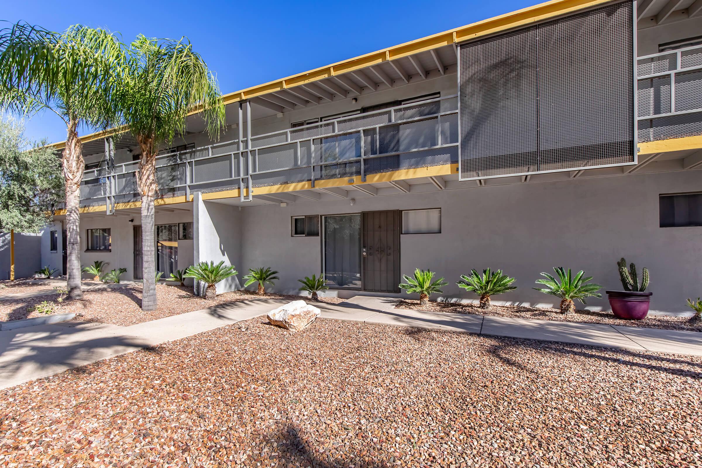 A view of a modern multi-unit building featuring a walkway with desert landscaping. The exterior is light grey with yellow accents. There are palm trees and succulents in the garden area, and the building has balconies with mesh screens. The sky is clear and blue, highlighting the sunny environment.