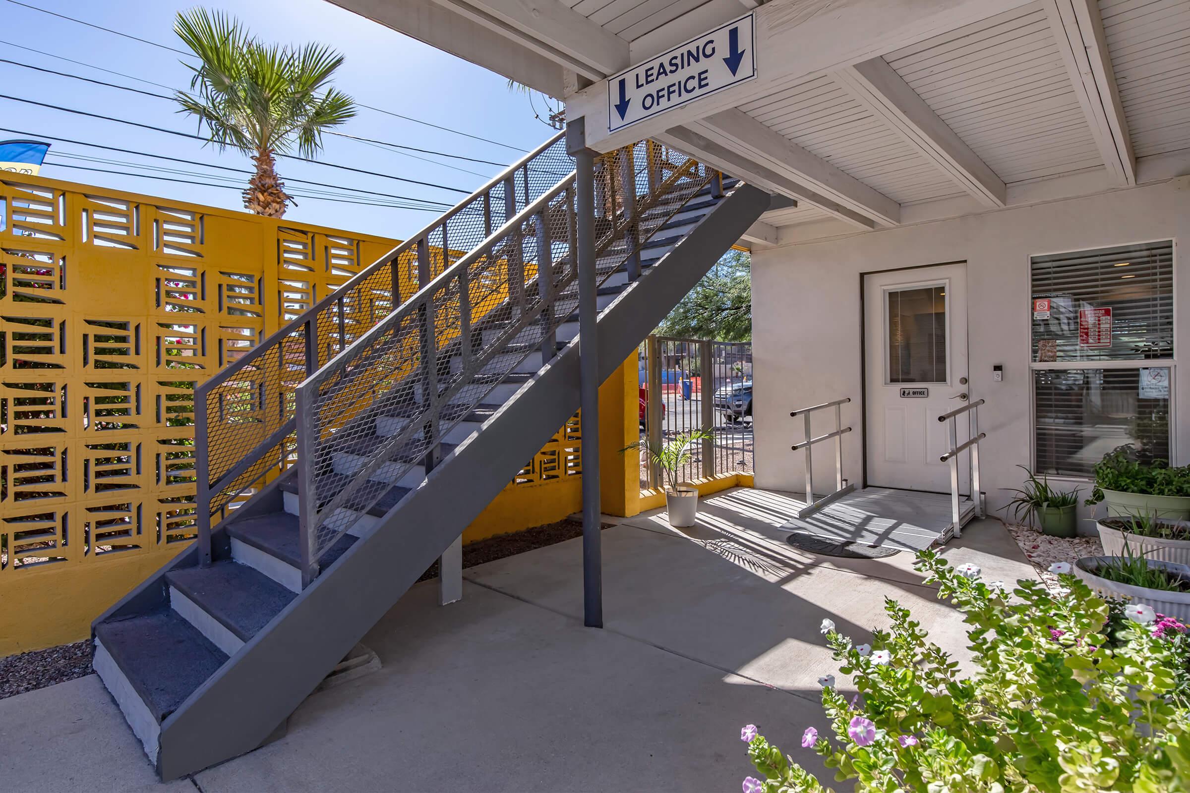 Exterior view of a leasing office with a staircase leading up to it. The office features a door with a sign indicating it's the leasing office, surrounded by potted plants. A bright yellow wall and a palm tree are visible, adding to the welcoming atmosphere. Sunlight casts shadows on the concrete.