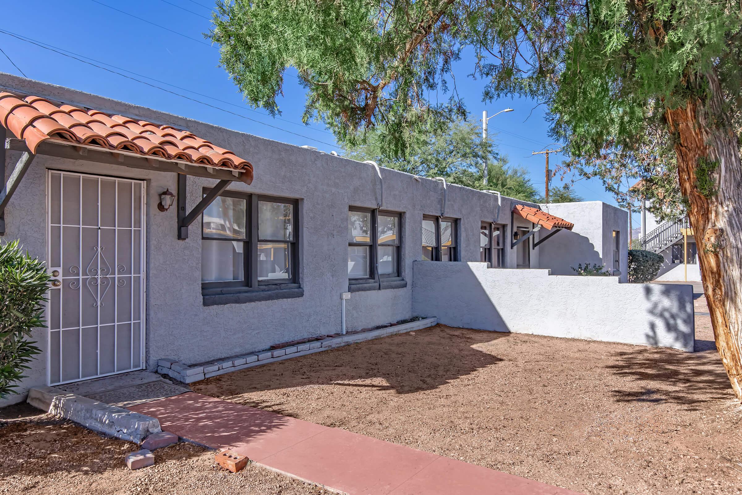 A single-story, grey stucco building with a red-tiled roof features a front porch and a decorative metal door. The landscape includes gravel and a few shrubs, with a clear blue sky above. The building has multiple windows, giving it a welcoming appearance.