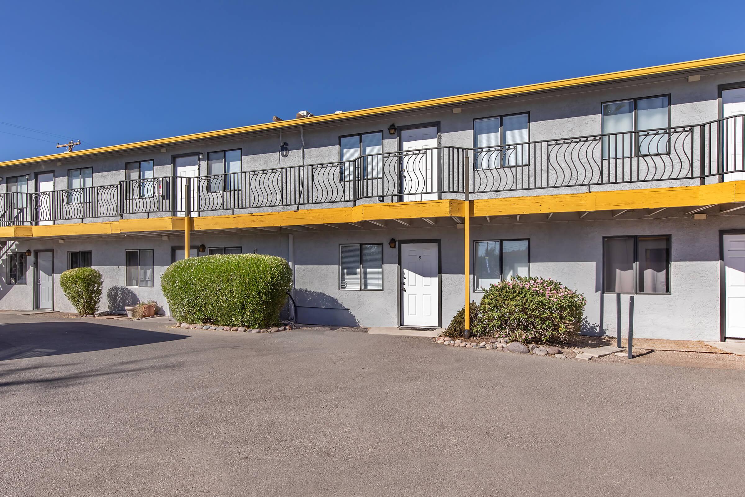 A two-story motel with a yellow overhang and gray exterior. The building features balconies with decorative railings, several doors, and a landscaped area with shrubs. The driveway is paved, and there is clear blue sky in the background.