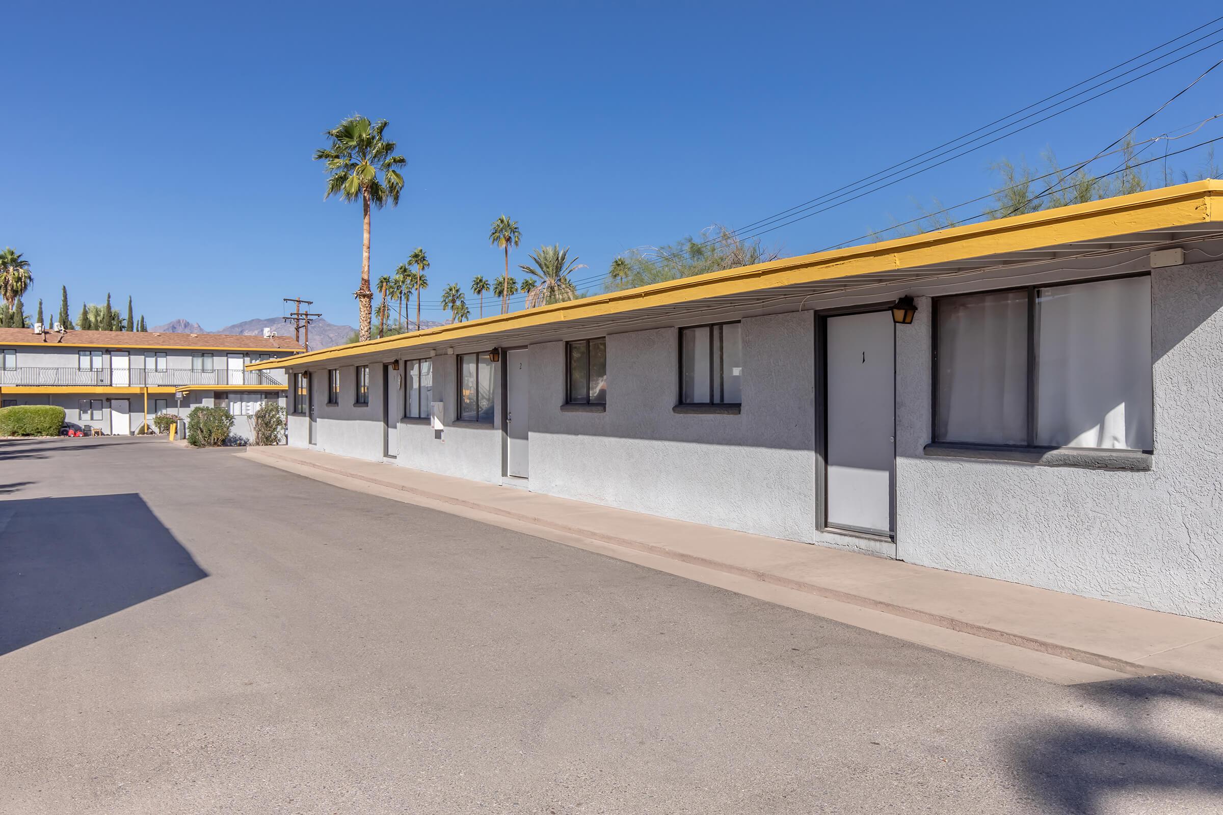 A view of a low-rise motel with a light-colored exterior and a yellow roof. Palm trees are visible in the background against a clear blue sky. The motel features multiple rooms with windows and doors facing a paved parking area.