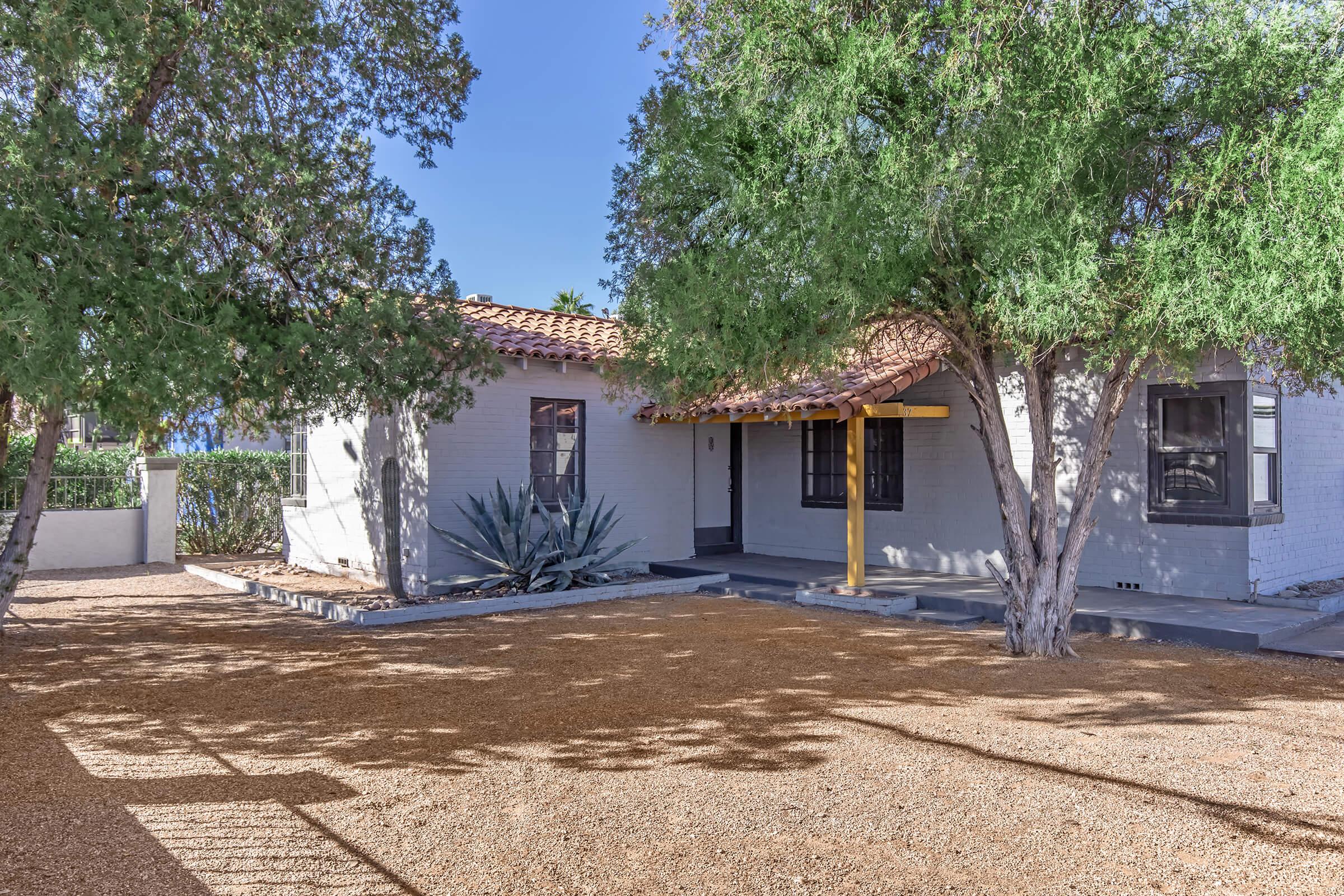 A cozy single-story house surrounded by trees, featuring a brown tiled roof, a wooden porch with yellow support beam, and a gravel yard. An agave plant is visible in the front, and the house has several windows that allow natural light. The setting is sunny and inviting.