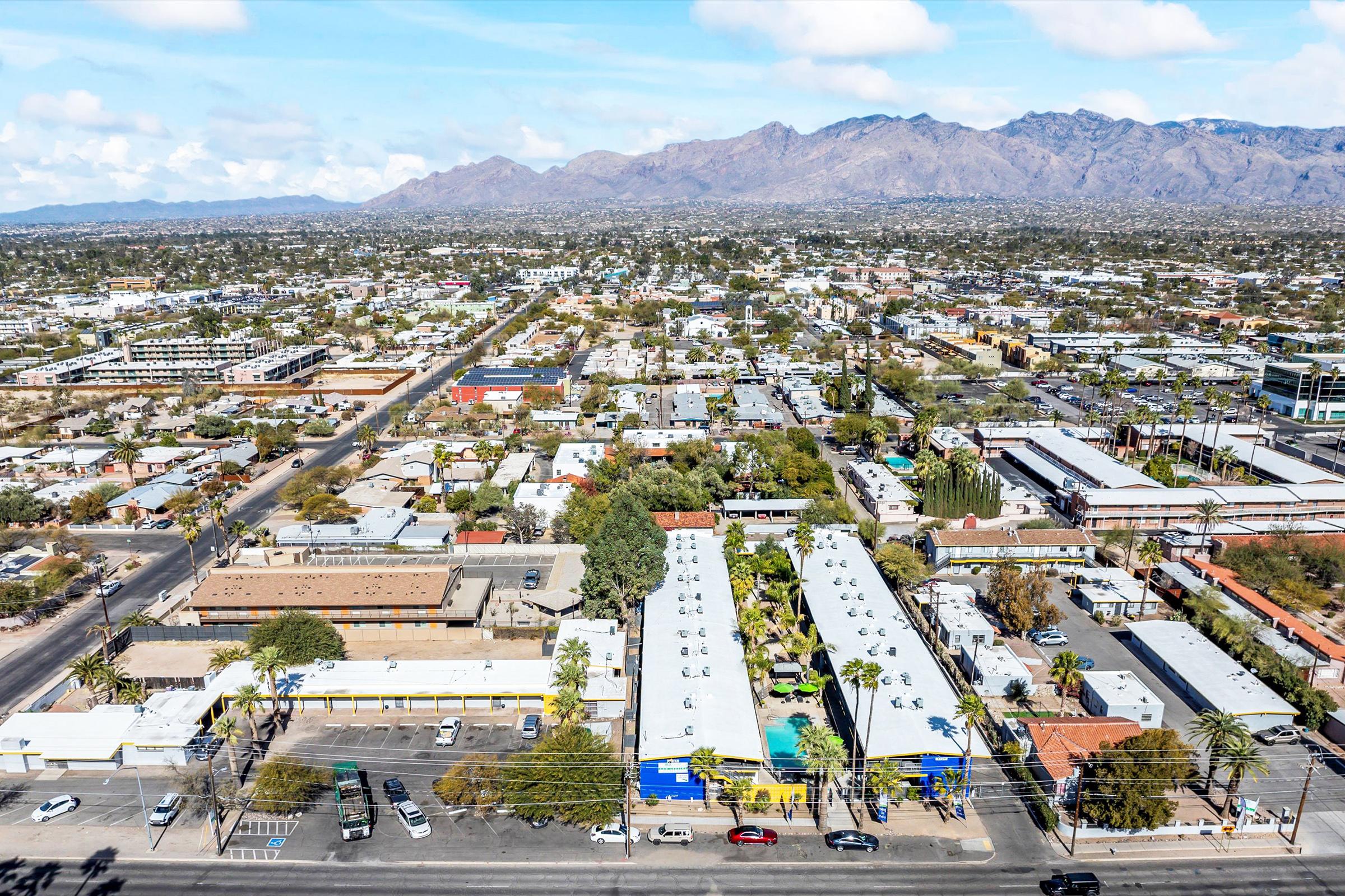 Aerial view of a city landscape featuring a mix of residential and commercial buildings, with streets running through the area. In the background, there are mountains and a clear blue sky, suggesting a sunny day. The greenery interspersed among the structures adds to the city's charm.