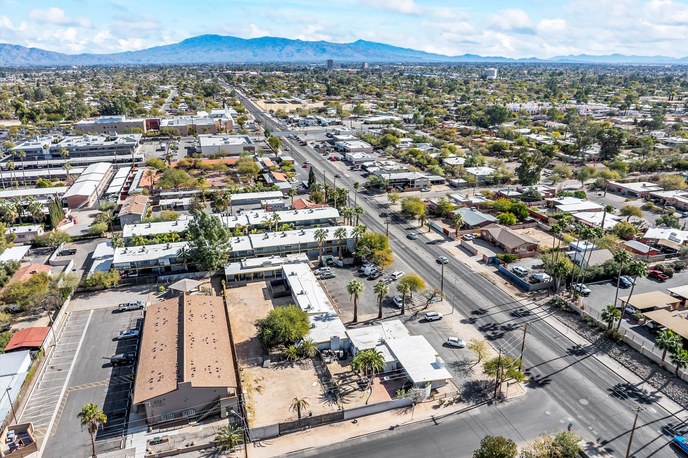 Aerial view of a suburban area with residential and commercial buildings, streets lined with palm trees, and distant mountains under a partly cloudy sky. The landscape features a mix of single-family homes and apartment complexes, with visible infrastructure and greenery throughout the area.