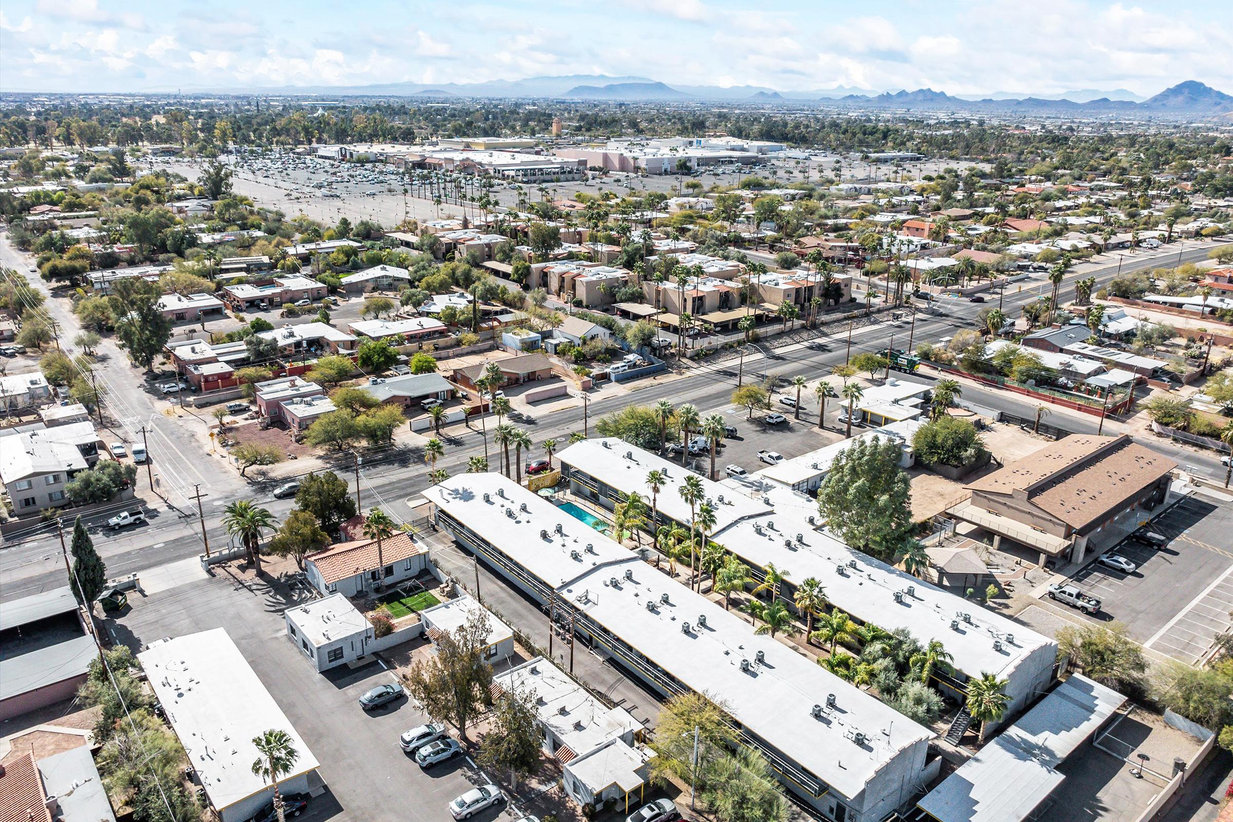 Aerial view of a suburban area featuring a mix of residential neighborhoods and commercial buildings. The image includes shops, parking lots, and a distant shopping center, with mountains visible in the background under a partly cloudy sky. Trees and roadways intersperse throughout the landscape.