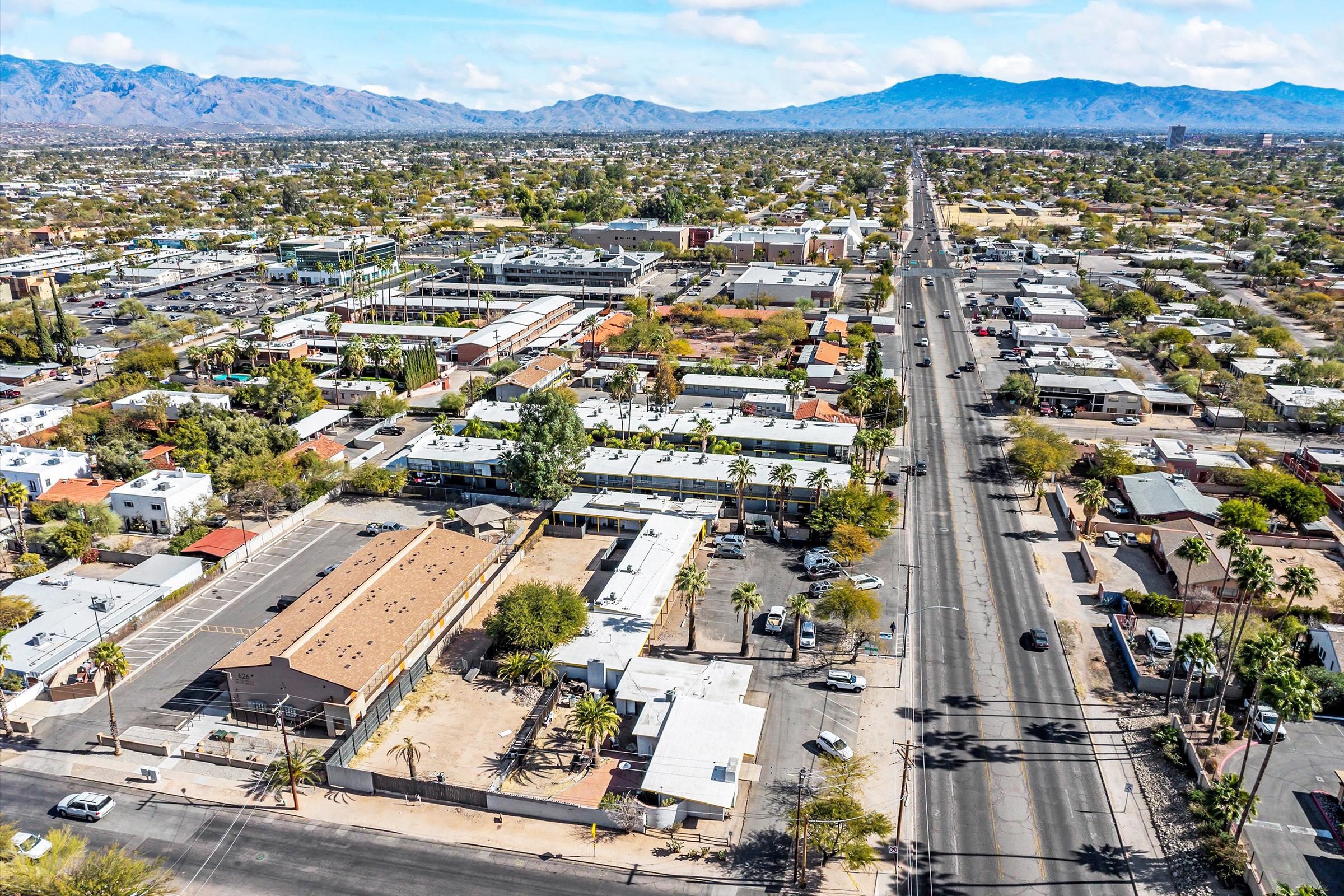 Aerial view of a suburban area featuring residential buildings, commercial establishments, and a wide road lined with palm trees. Mountains are visible in the background, and various vehicles are traveling along the road. The scene captures a vibrant community landscape with a mix of buildings and greenery.