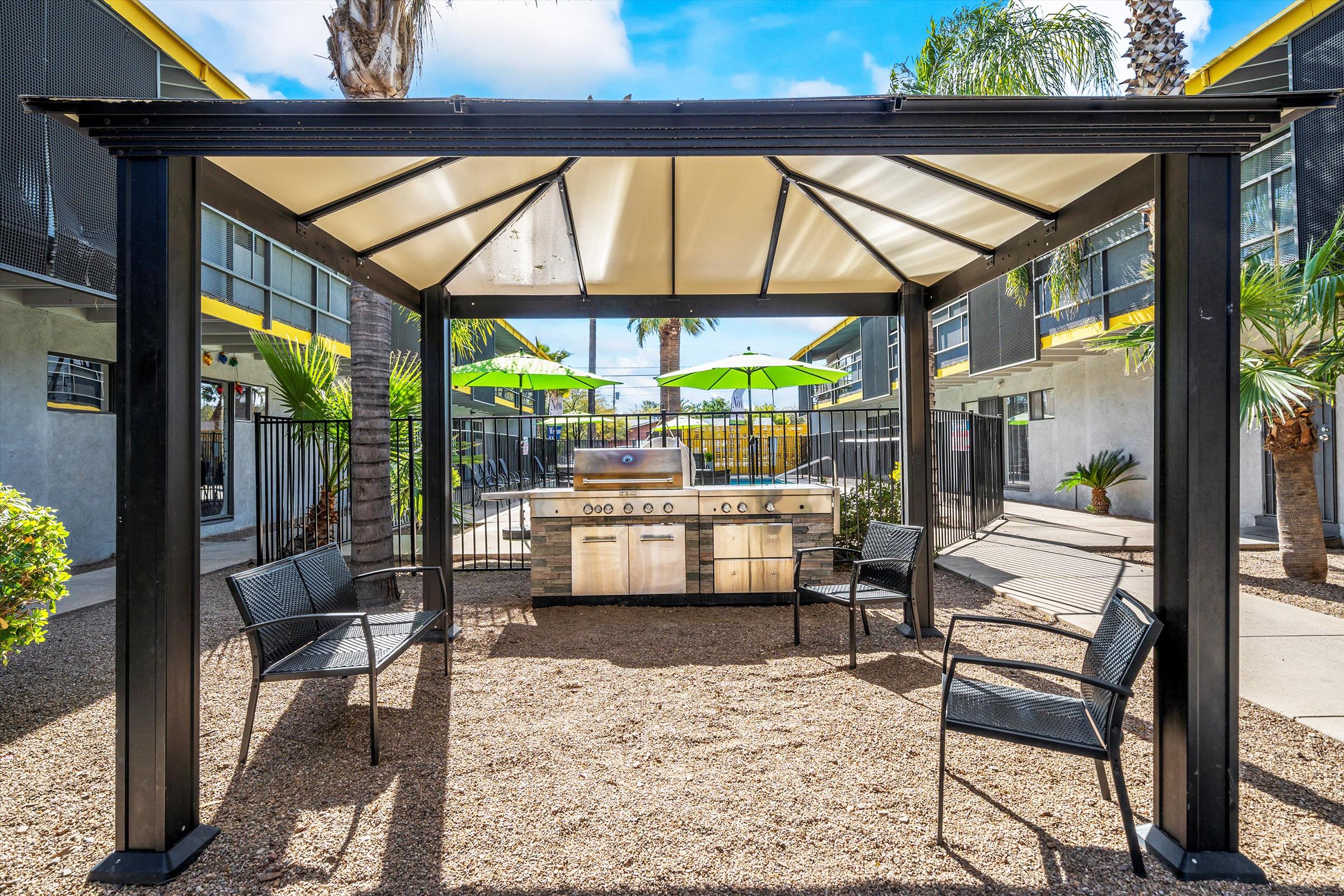 Outdoor grilling area covered by a pergola, featuring a stainless steel barbecue grill and four black chairs. Surrounded by landscaping with palm trees and green umbrellas, with an apartment complex in the background under a bright blue sky.
