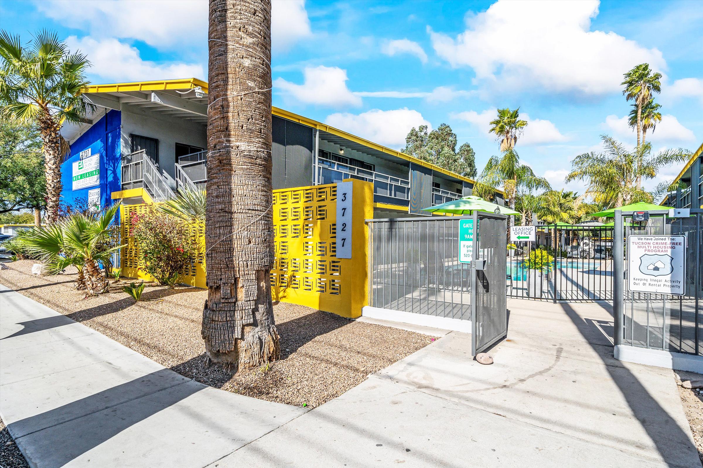 A modern building with a blue and yellow exterior, featuring yellow lockers and a gated entrance. Palm trees and greenery surround the area, with a swimming pool visible in the background. Clear sky with fluffy clouds enhances the vibrant atmosphere.