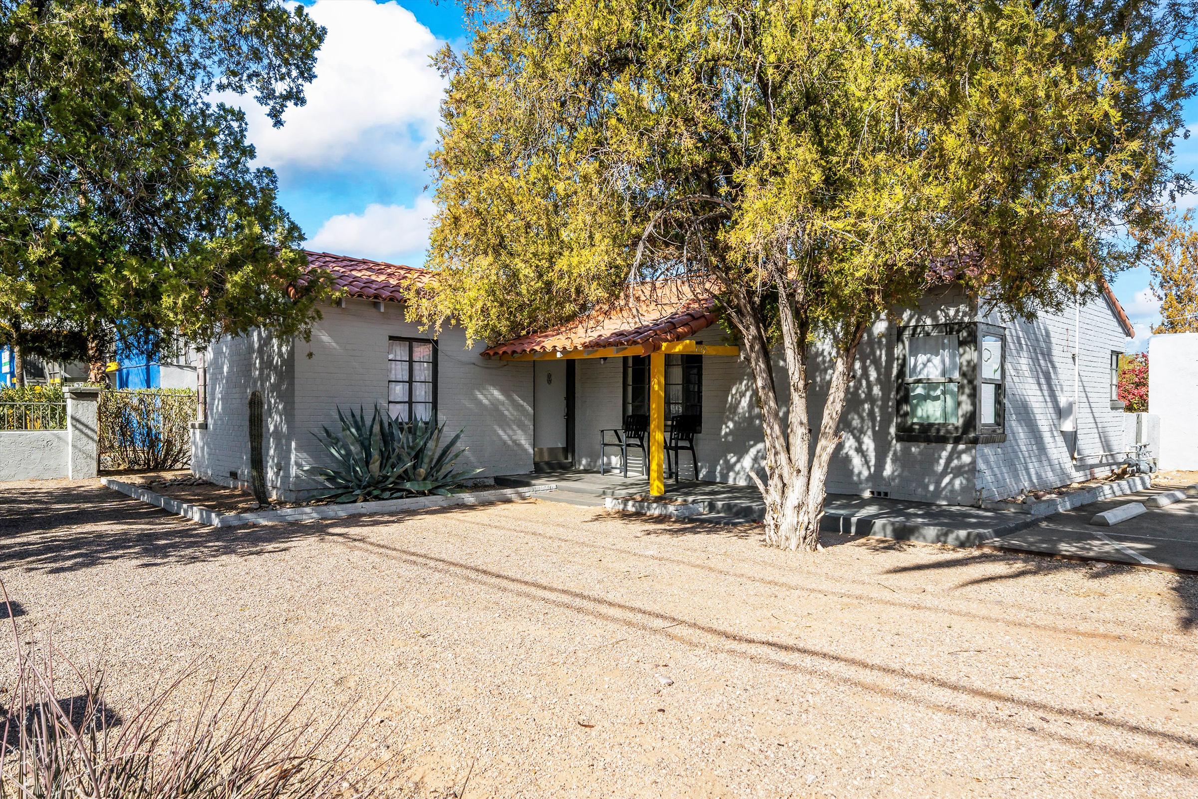 A charming single-story home with a tiled roof, surrounded by trees and shrubs. The house features a front porch with yellow columns, and a dirt driveway leading up to it. Blue skies with a few clouds are visible in the background, giving the scene a bright, welcoming atmosphere.