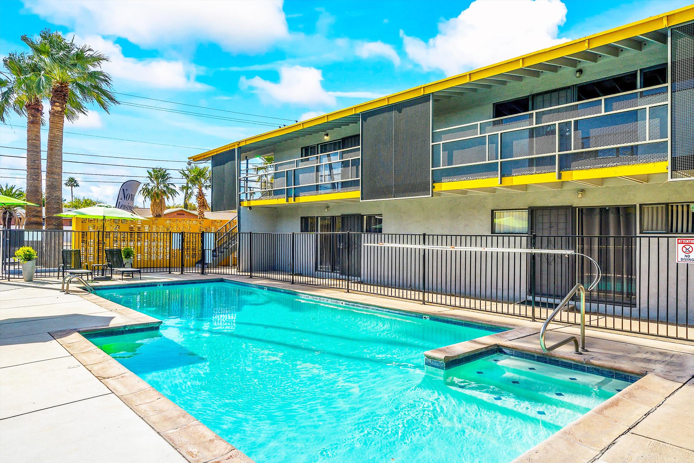A bright and inviting swimming pool area in front of a modern, two-story apartment building with yellow accents. Palm trees are visible, and the sky is clear with a few clouds. A fenced patio area surrounds the pool, enhancing the relaxing atmosphere.