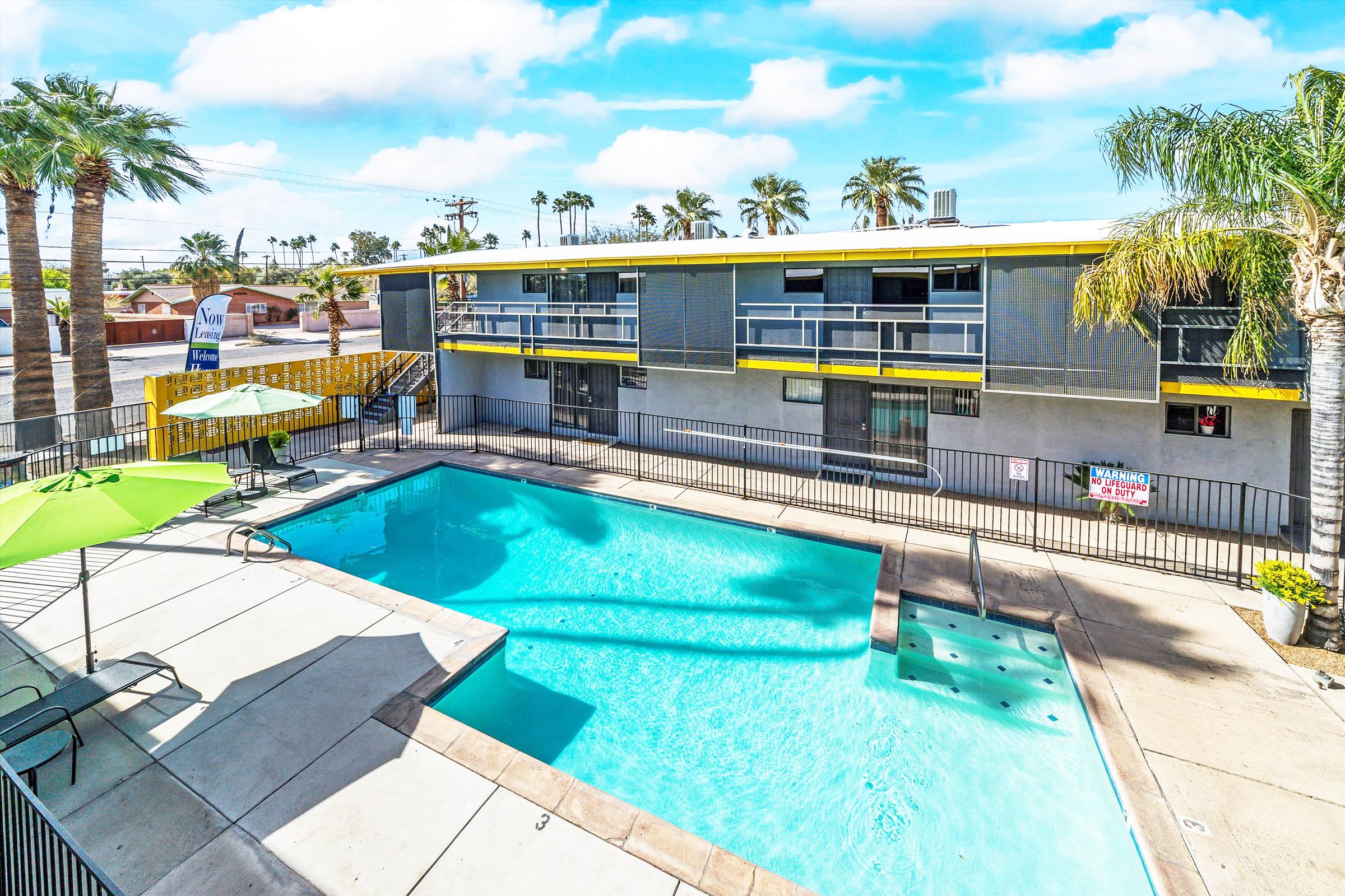 A bright and inviting outdoor area featuring a swimming pool surrounded by palm trees. The modern building has a yellow accent and balconies, with lounge chairs and umbrellas by the poolside. Clear blue skies add to the cheerful atmosphere.