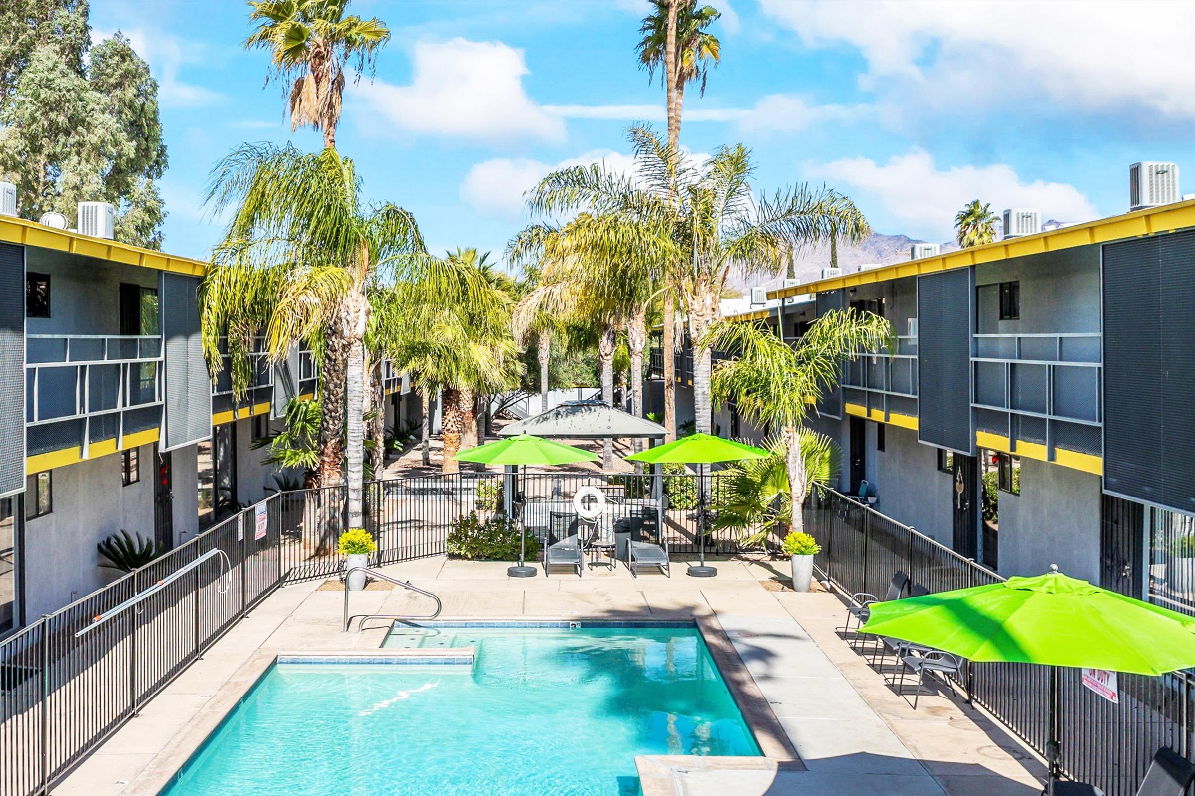 A vibrant outdoor swimming pool surrounded by palm trees and lounge chairs. Two-story buildings with balconies flank the pool area, while large green umbrellas provide shade. The scene is bright and inviting under a partly cloudy blue sky.