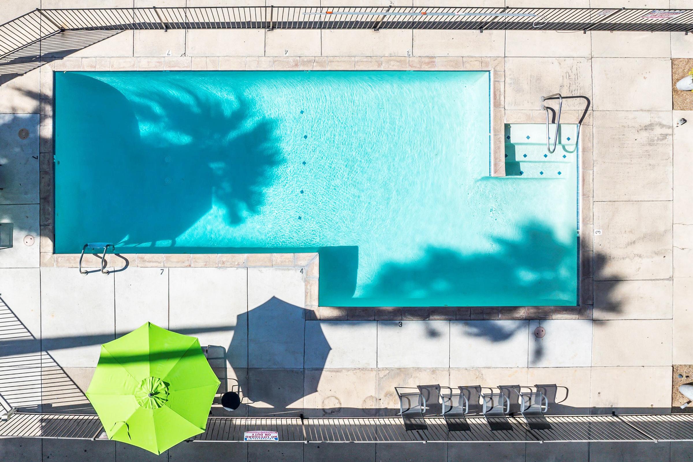 Aerial view of a swimming pool with clear blue water, surrounded by concrete decking. There are lounge chairs next to the pool and two umbrellas—one green and one white. Shadows from palm trees rest on the water surface, creating a relaxing atmosphere.