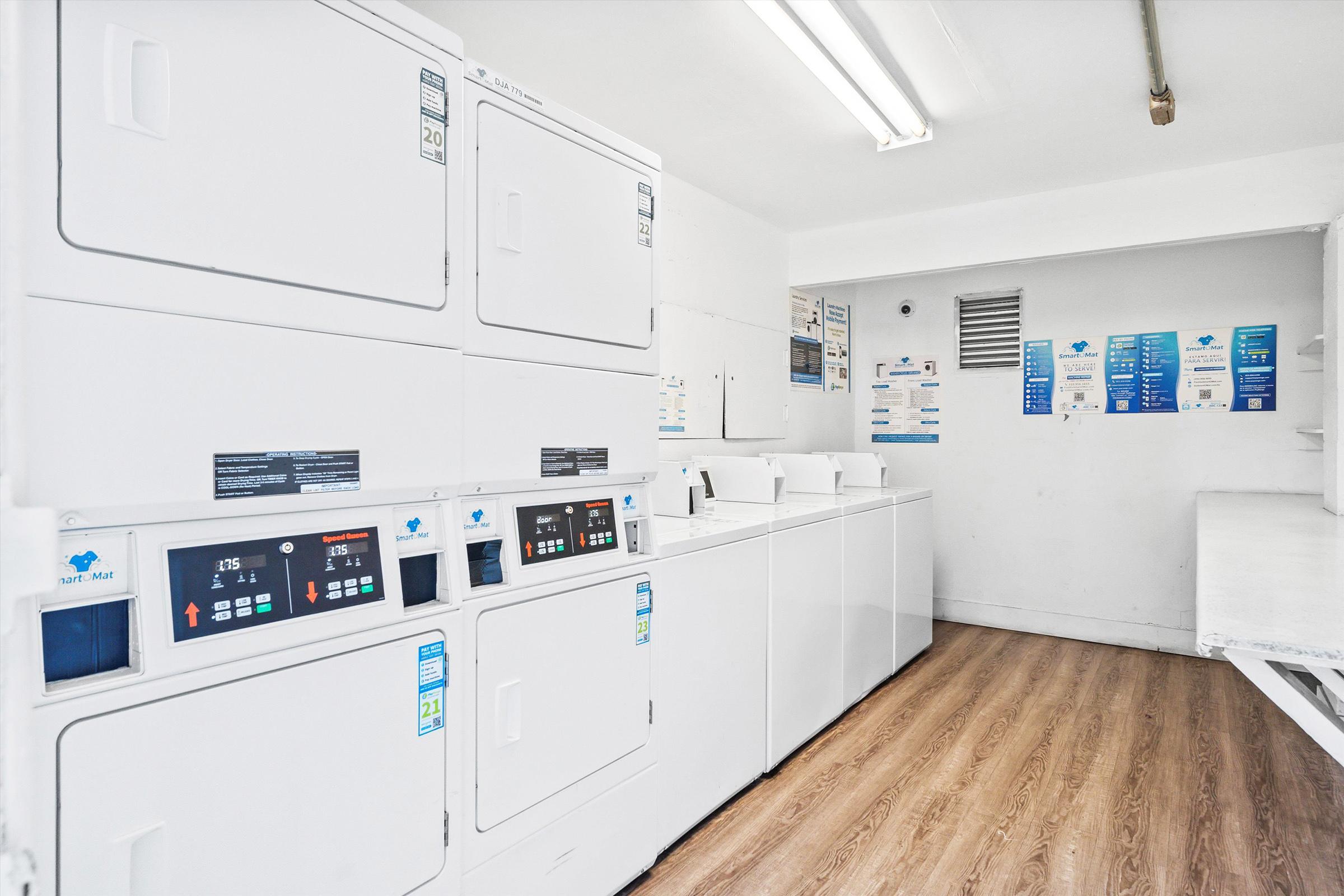 A clean laundry room featuring multiple white washing machines and dryers. The machines are arranged in a row along one wall, with digital displays and buttons visible. The floor is wooden, and there are informational signs on the walls, indicating laundry instructions and prices. The lighting is bright and even.