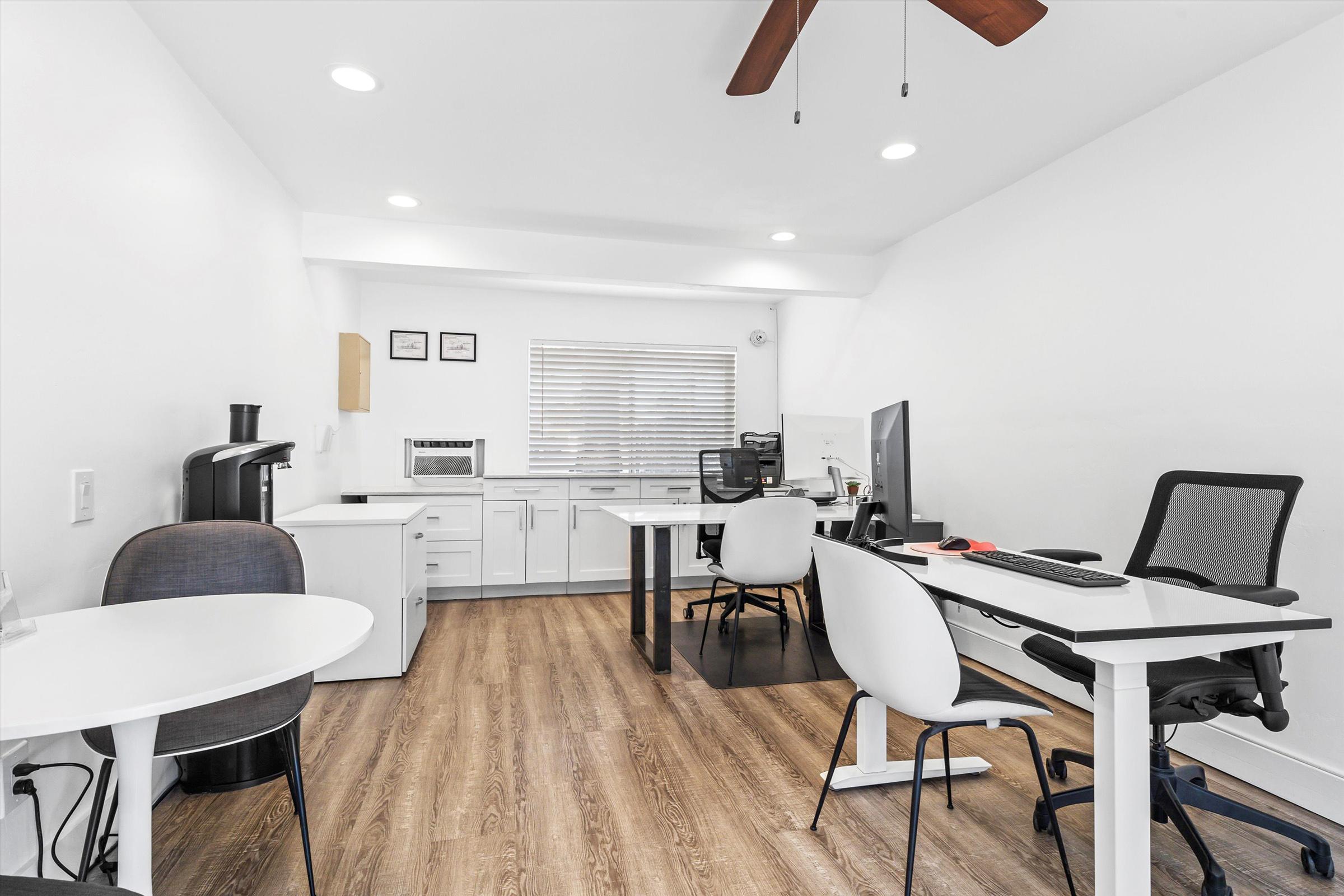 Modern office interior featuring white walls and wooden flooring. There are two workstations with chairs, a desk with a computer, and a small round table. A coffee machine and microwave are visible. Large windows allow natural light, and there are ceiling lights and a ceiling fan, creating a bright and open atmosphere.