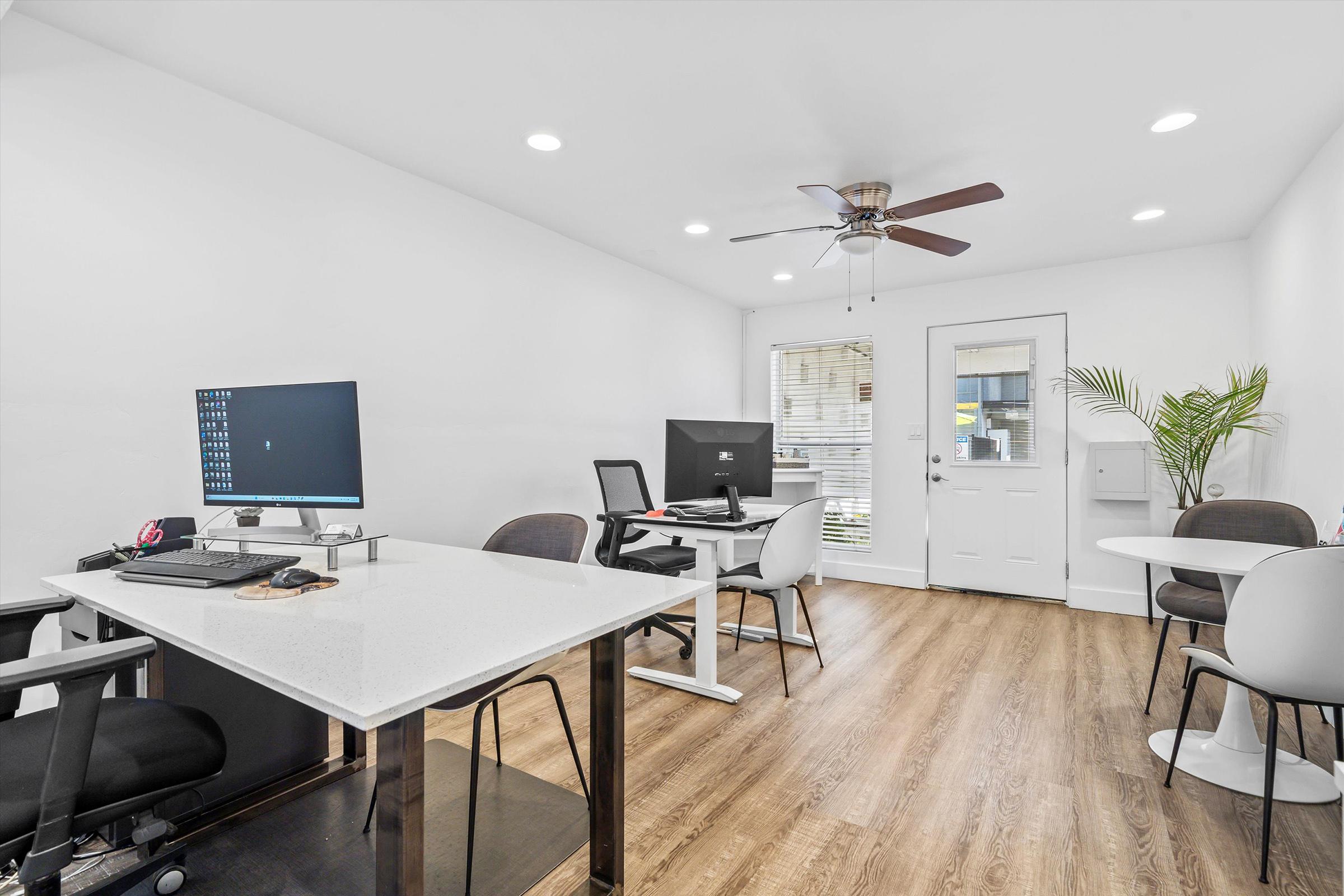 A modern office space featuring multiple workstations. There is a desk with a computer and a chair, a second workstation with another computer, and a door leading outside. Natural light enters through a window, complemented by a ceiling fan and a small plant in the corner. The flooring is wooden.