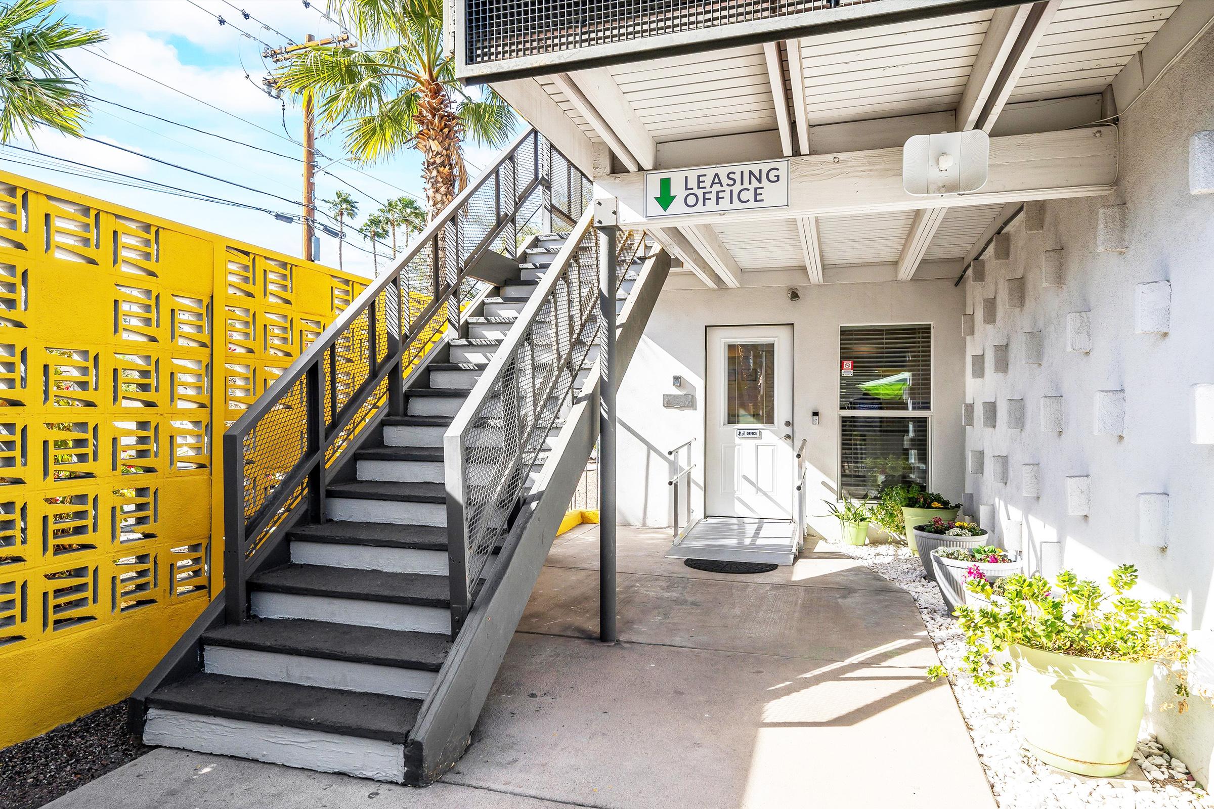 Exterior view of a leasing office with a staircase leading up to the entrance. The building features a gray facade and a vibrant yellow wall with decorative patterns. Planters with greenery are placed along the pathway. Bright sunlight illuminates the scene, highlighting the inviting atmosphere.