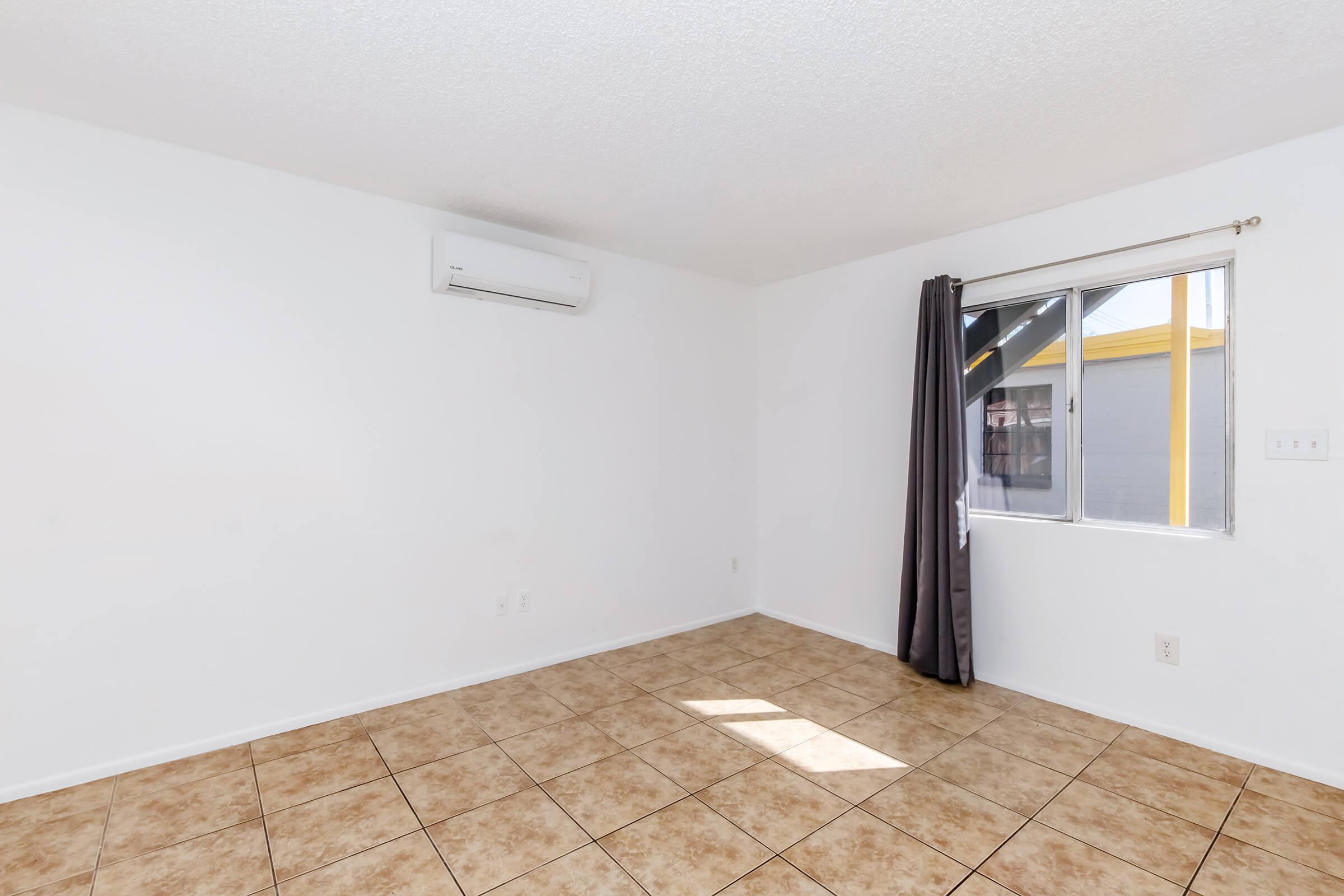 A simple, empty room featuring tiled flooring, a window with a curtain, and a wall-mounted air conditioning unit. The walls are painted white, and there is ample natural light coming through the window.